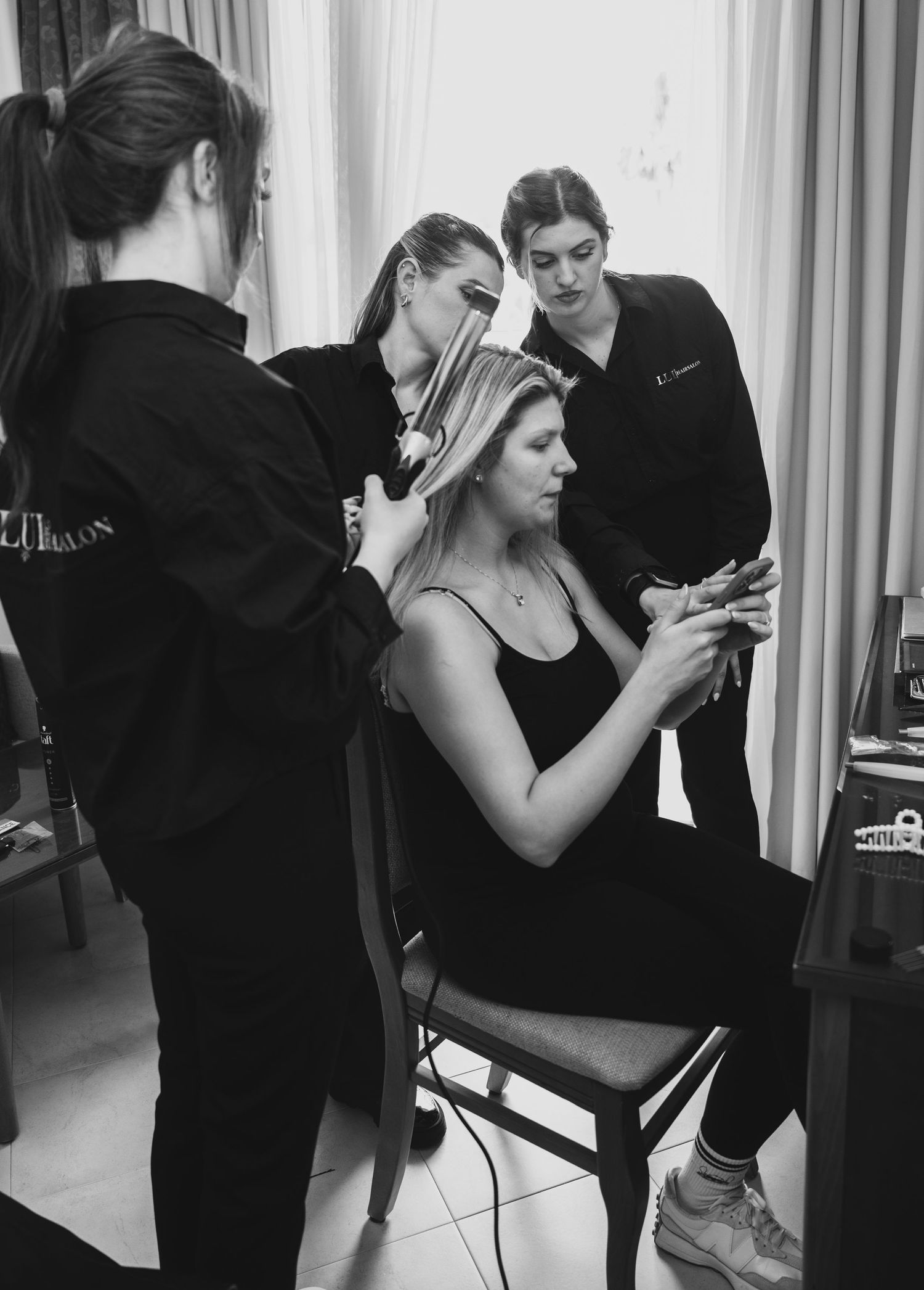 Black and white photo of bride seated in chair getting hair done by two stylists.