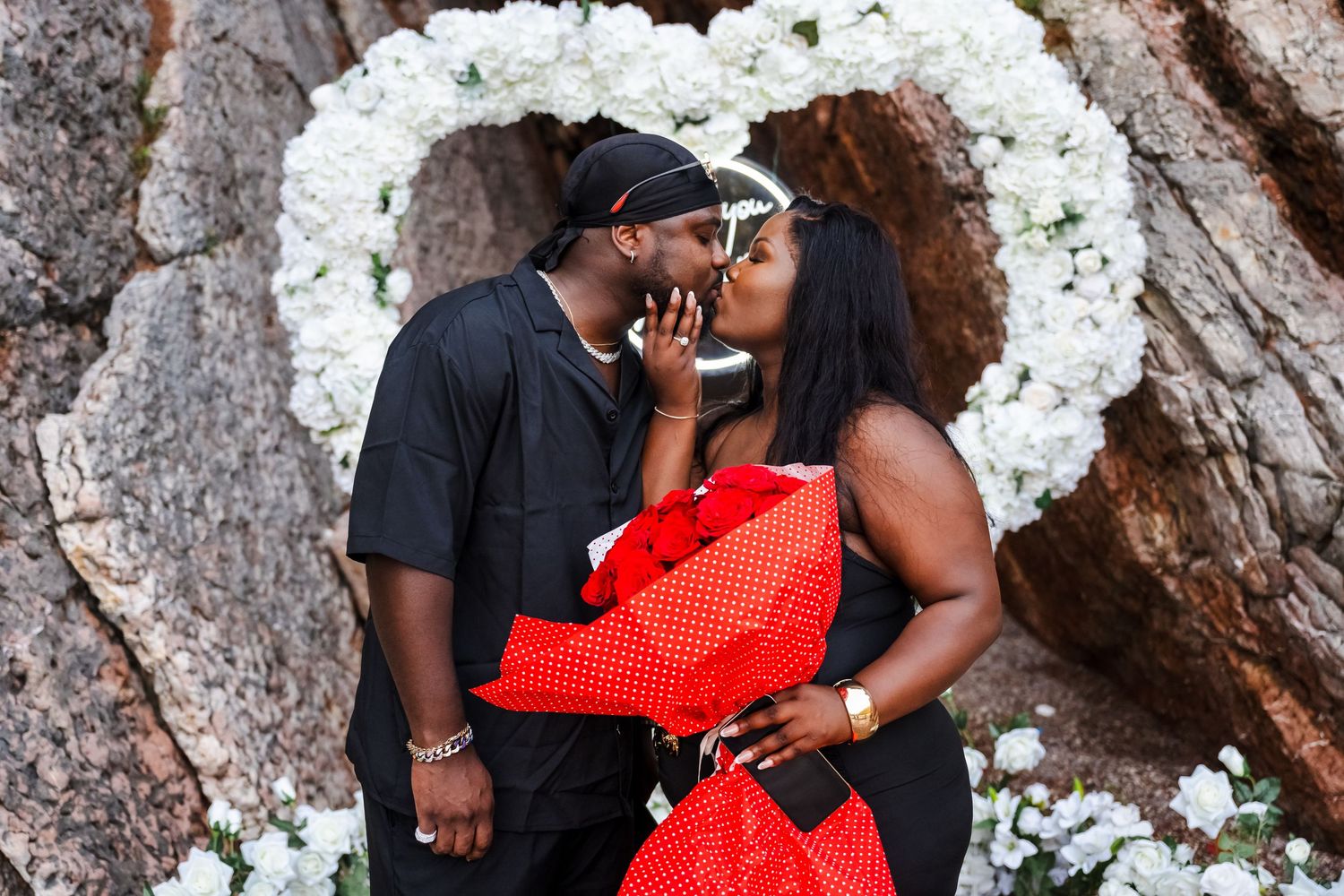 Happy couple kisses in front of white floral heart arch during romantic outdoor marriage proposal.