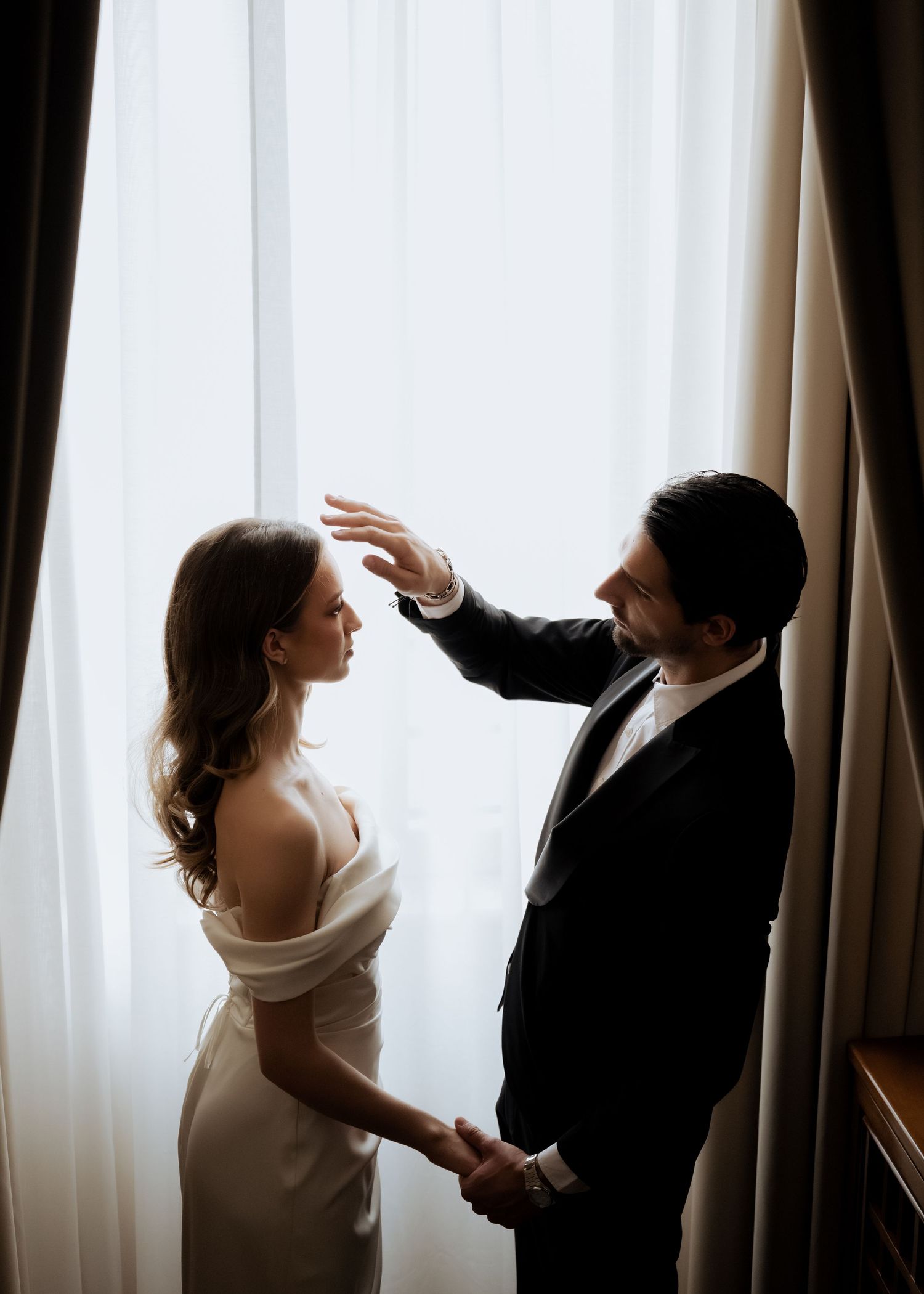 Groom gently raises hand over bride near sunlit curtained window in romantic wedding photo.