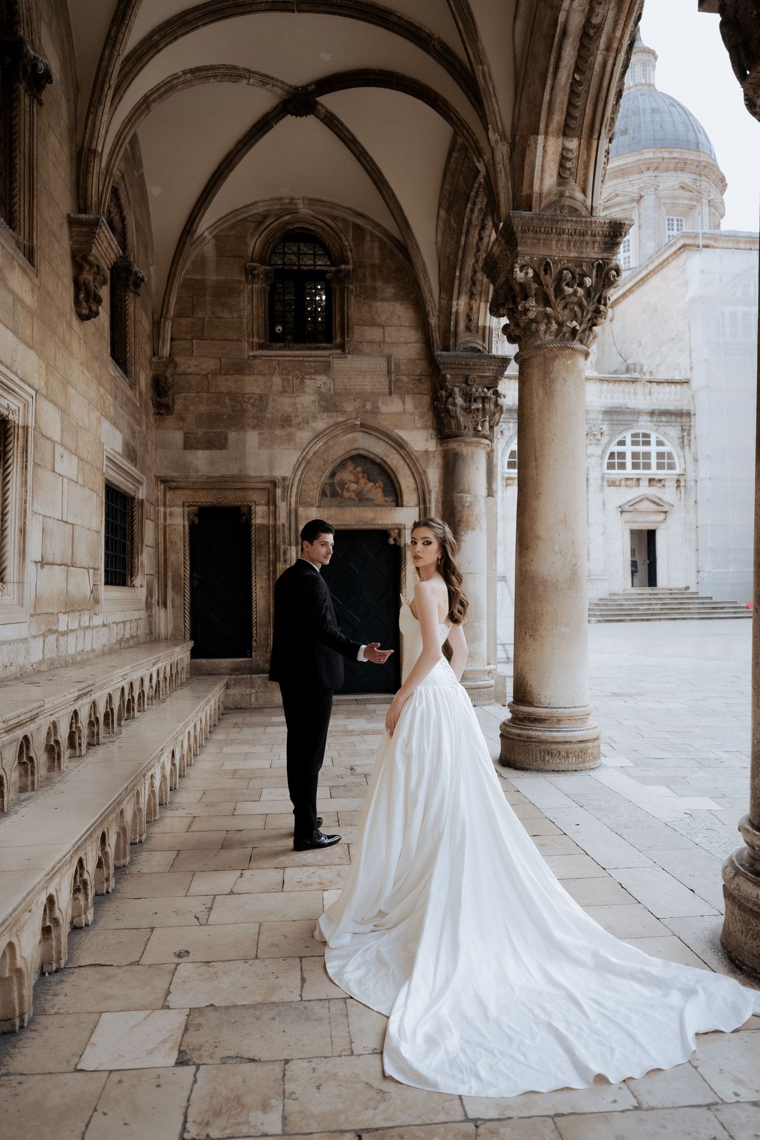 Bride in a flowing white gown with long train stands beside groom in a vaulted stone arcade.