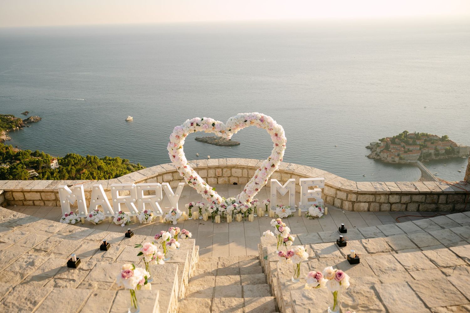 Aerial view of a romantic Marry Me proposal setup with floral heart arch overlooking the ocean.