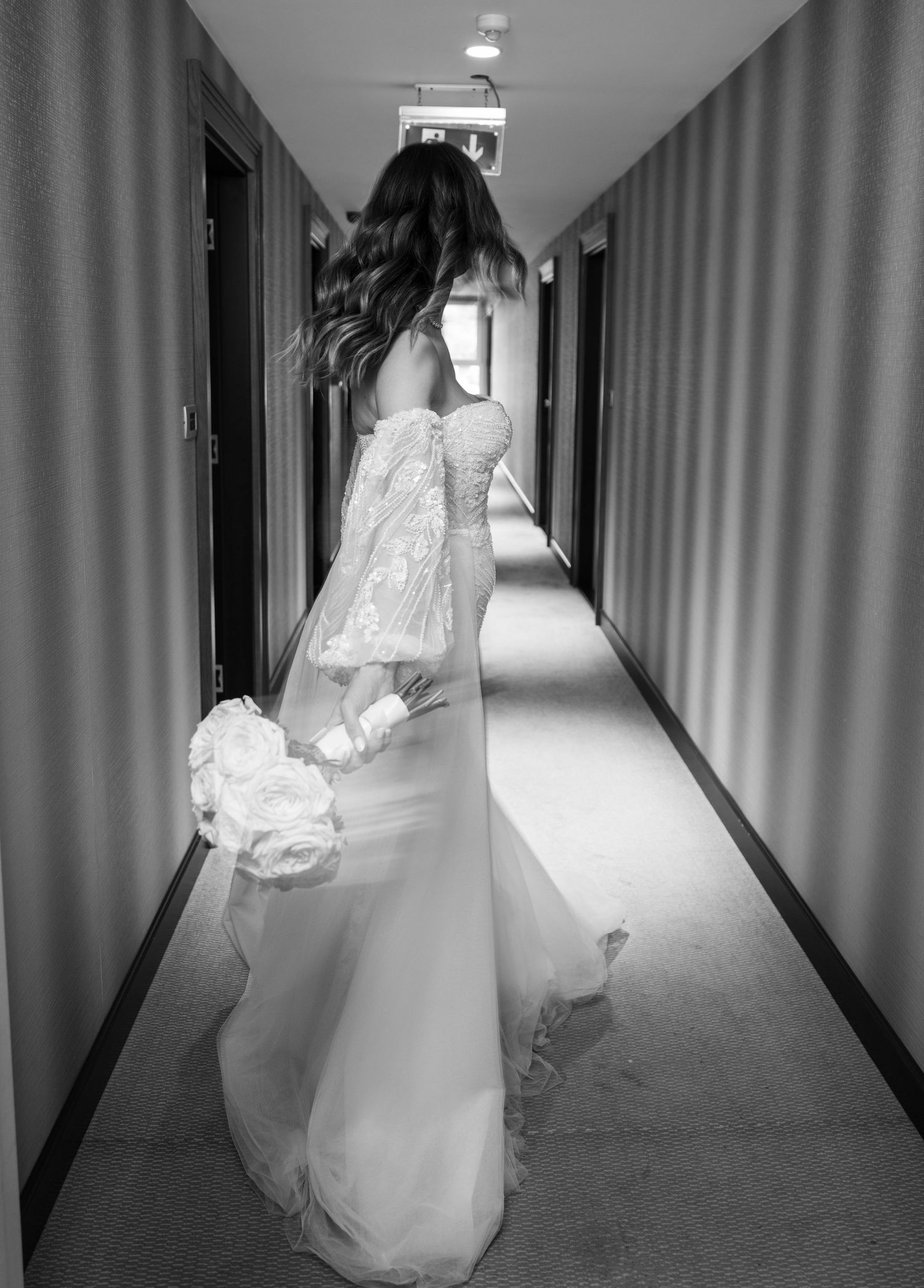 Black and white photo of bride in lace wedding dress walking down hotel corridor holding white bouquet.