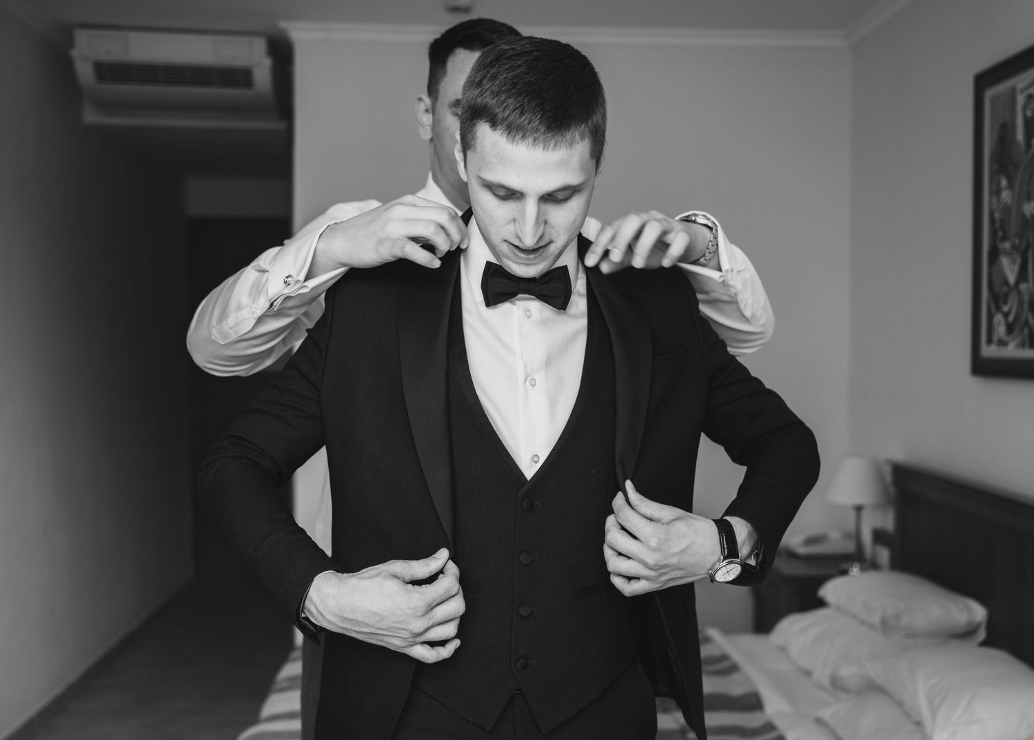 Black and white photo of a groom in a tuxedo being helped with his jacket by a groomsman.