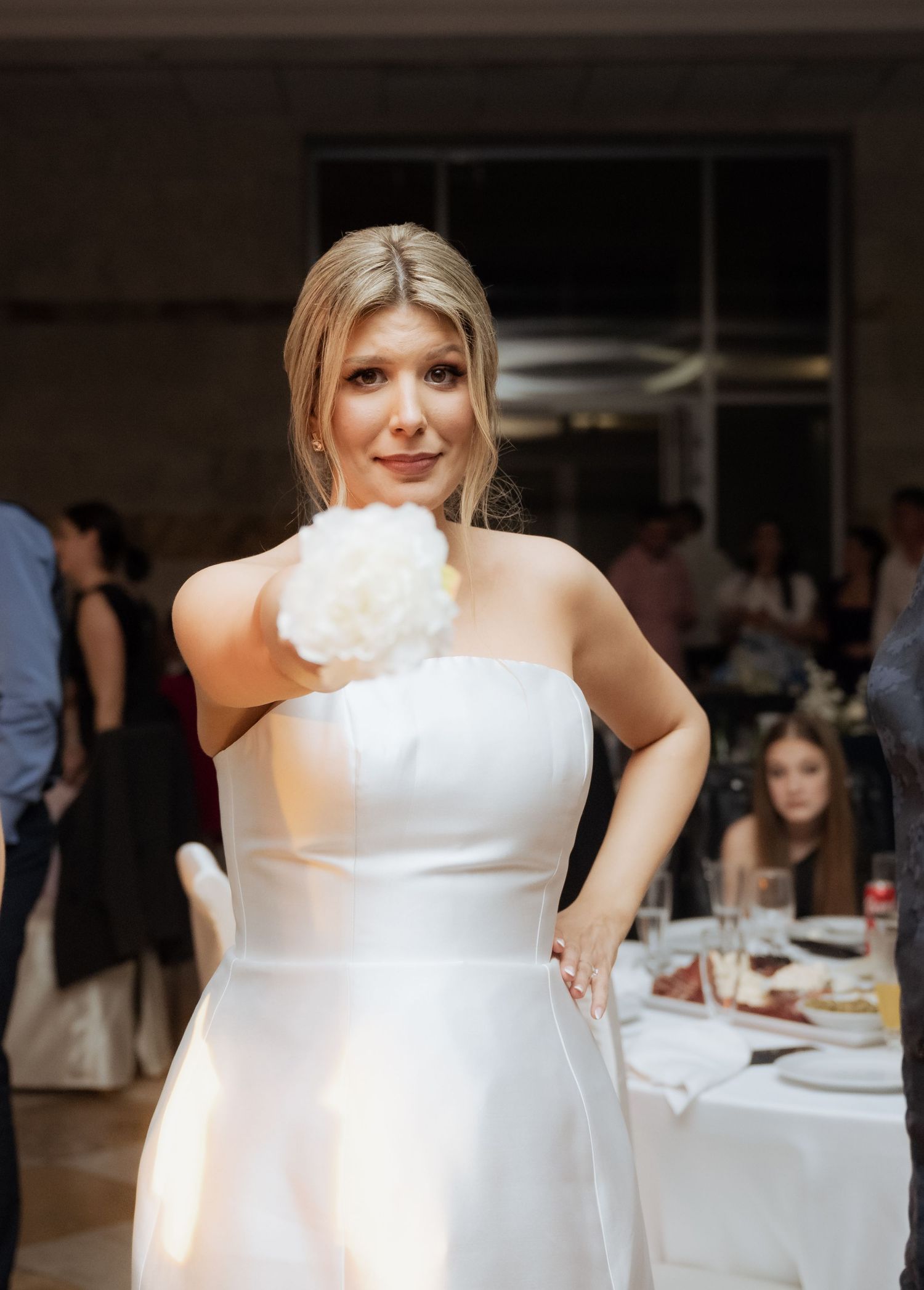 Elegant blonde bride in strapless white gown holding white floral bouquet at wedding reception.