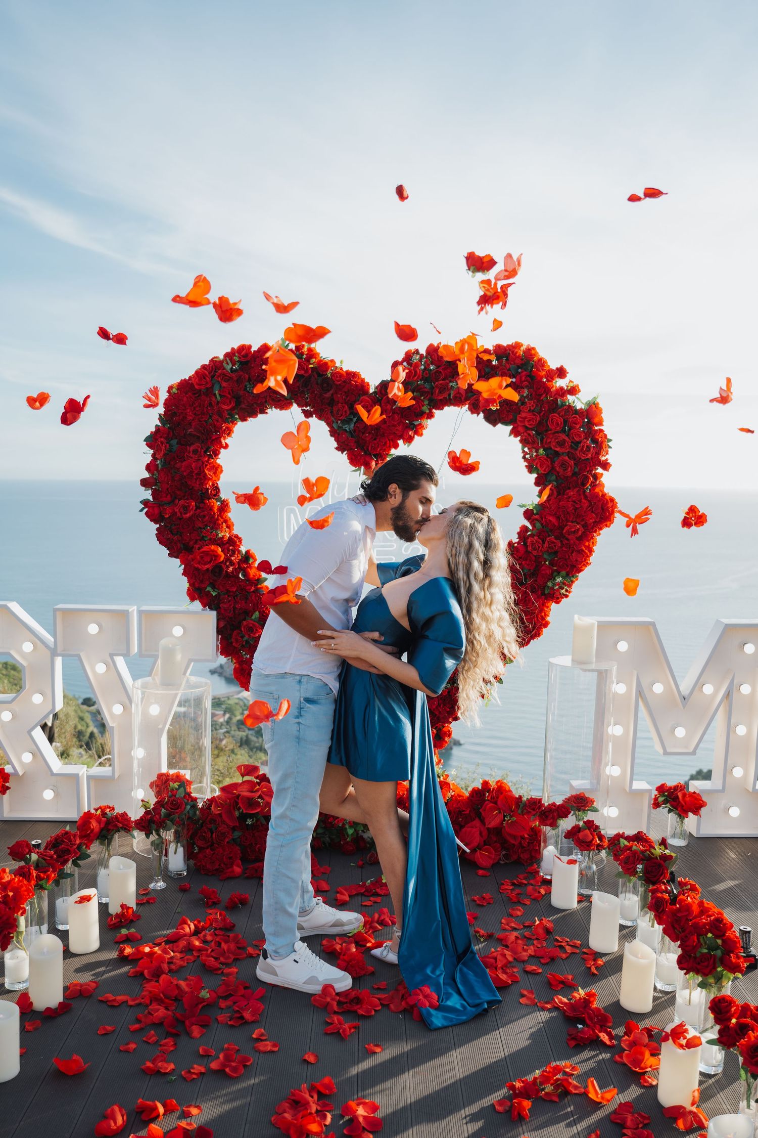 Couple kissing at romantic marriage proposal setup with red rose heart arch and rose petals.