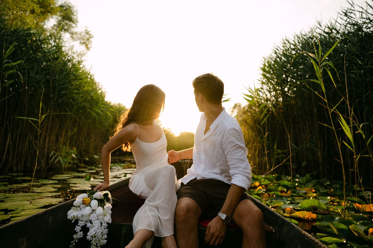 Couple sitting together in a boat among lily pads with golden sunlight filtering through reeds.