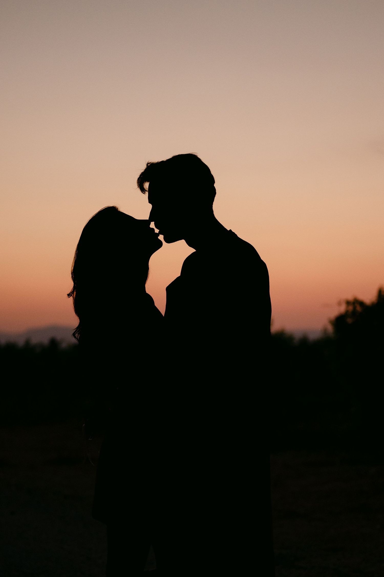 Romantic silhouette of couple almost kissing against vibrant orange and pink sunset sky.