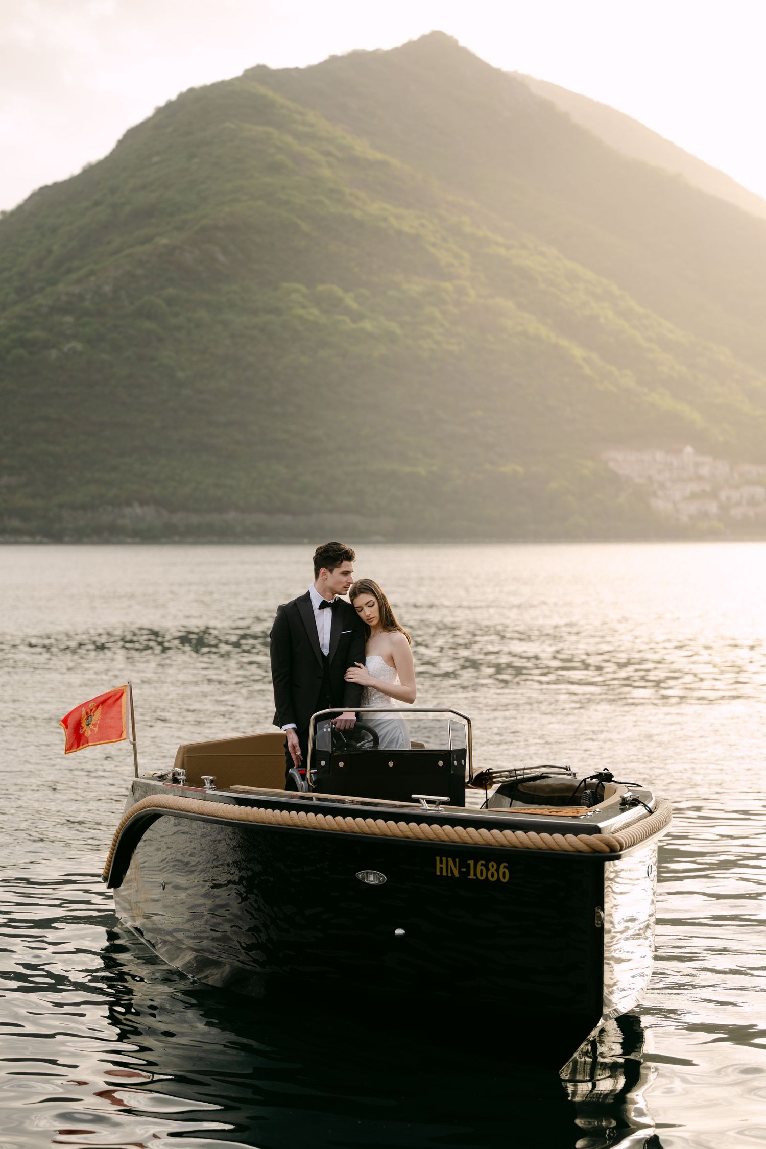 Bride and groom standing on a sleek black boat with Montenegro flag, framed by a dramatic mountain sunset.