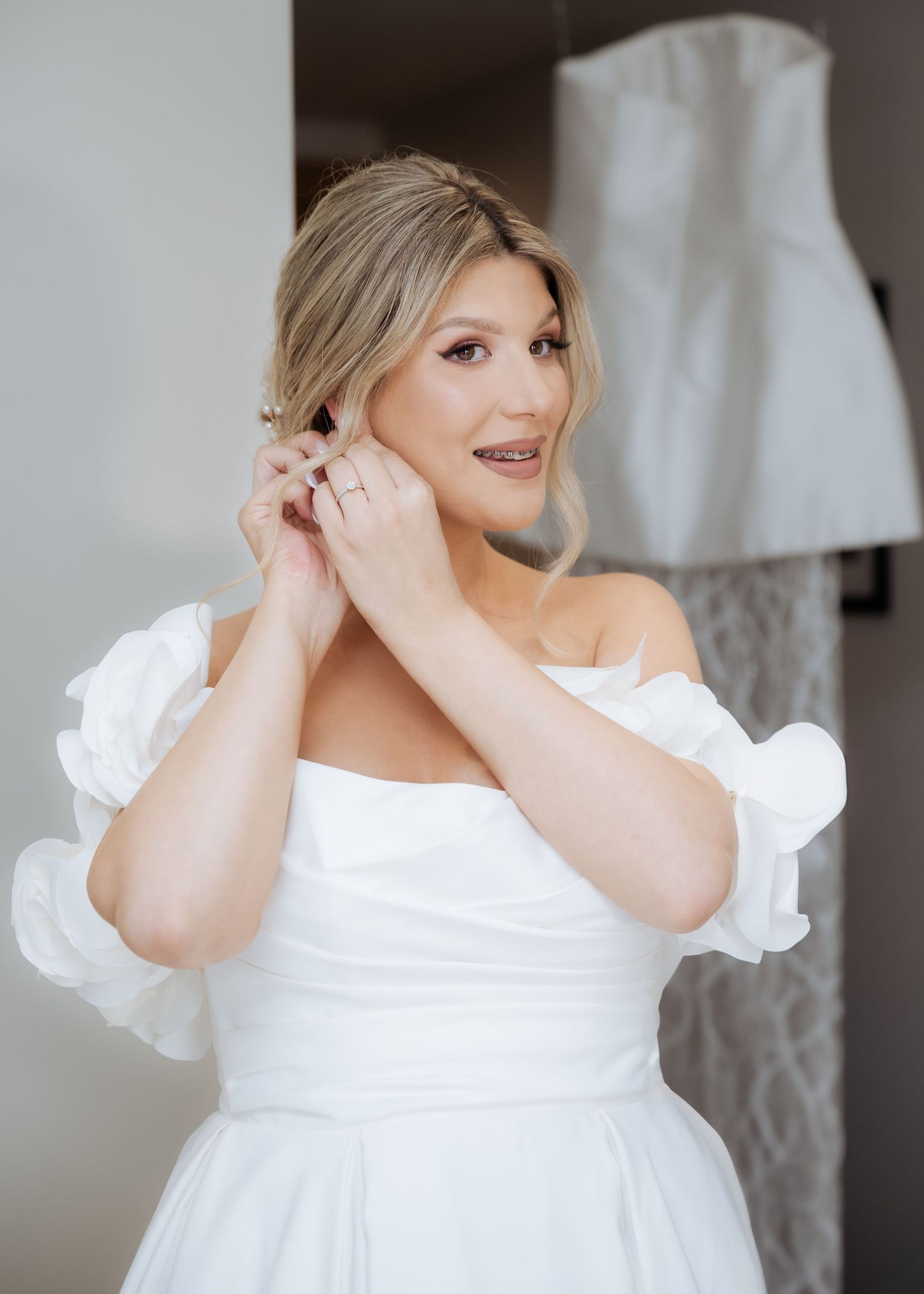 Beautiful blonde bride in off-shoulder white wedding dress adjusting earrings before ceremony.