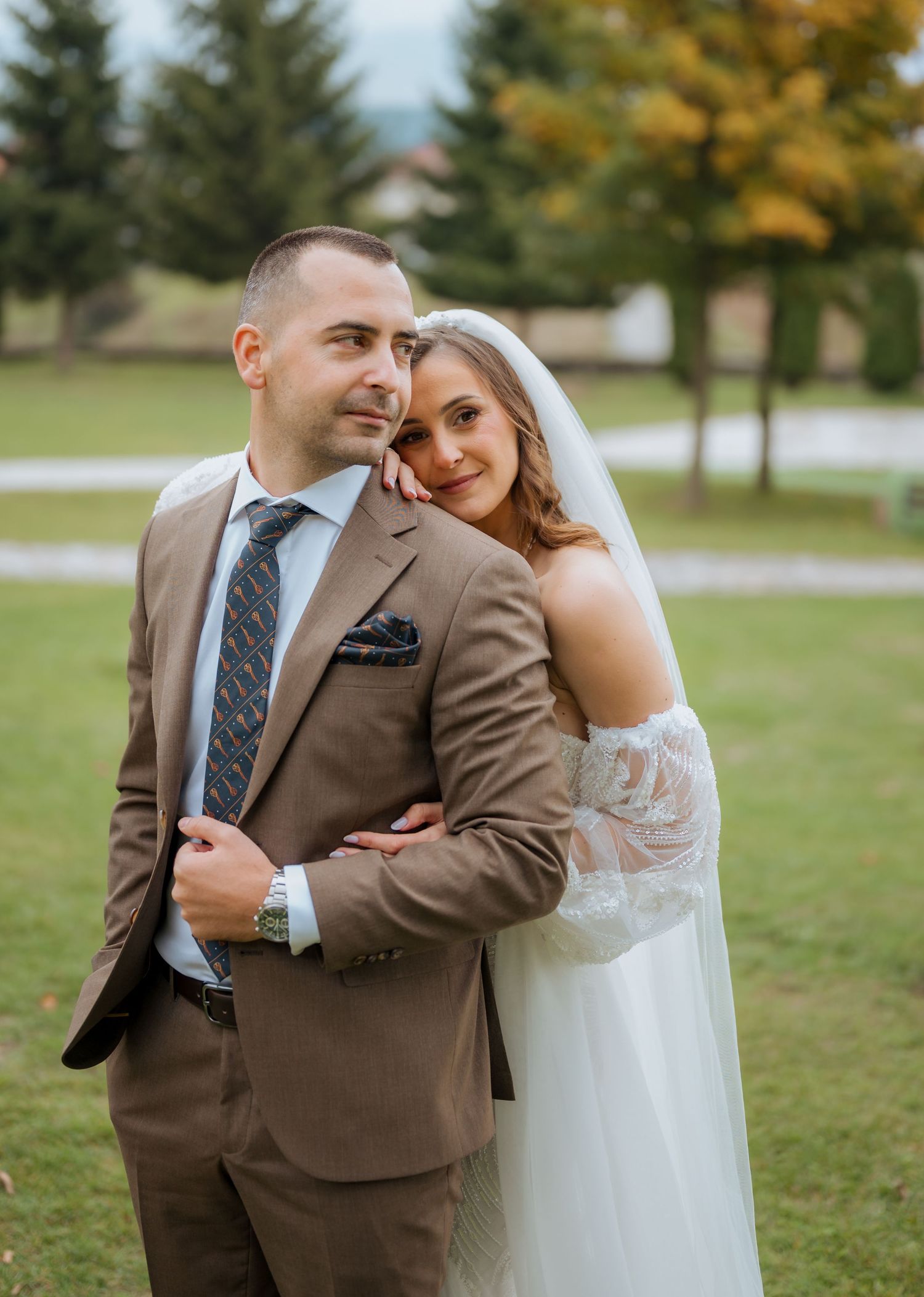 Bride in off-shoulder lace wedding dress leans on groom in brown suit outdoors with green trees in background.