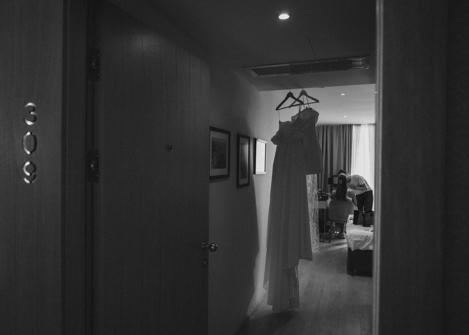 Black and white photo of a white wedding dress hanging in a hotel room doorway.
