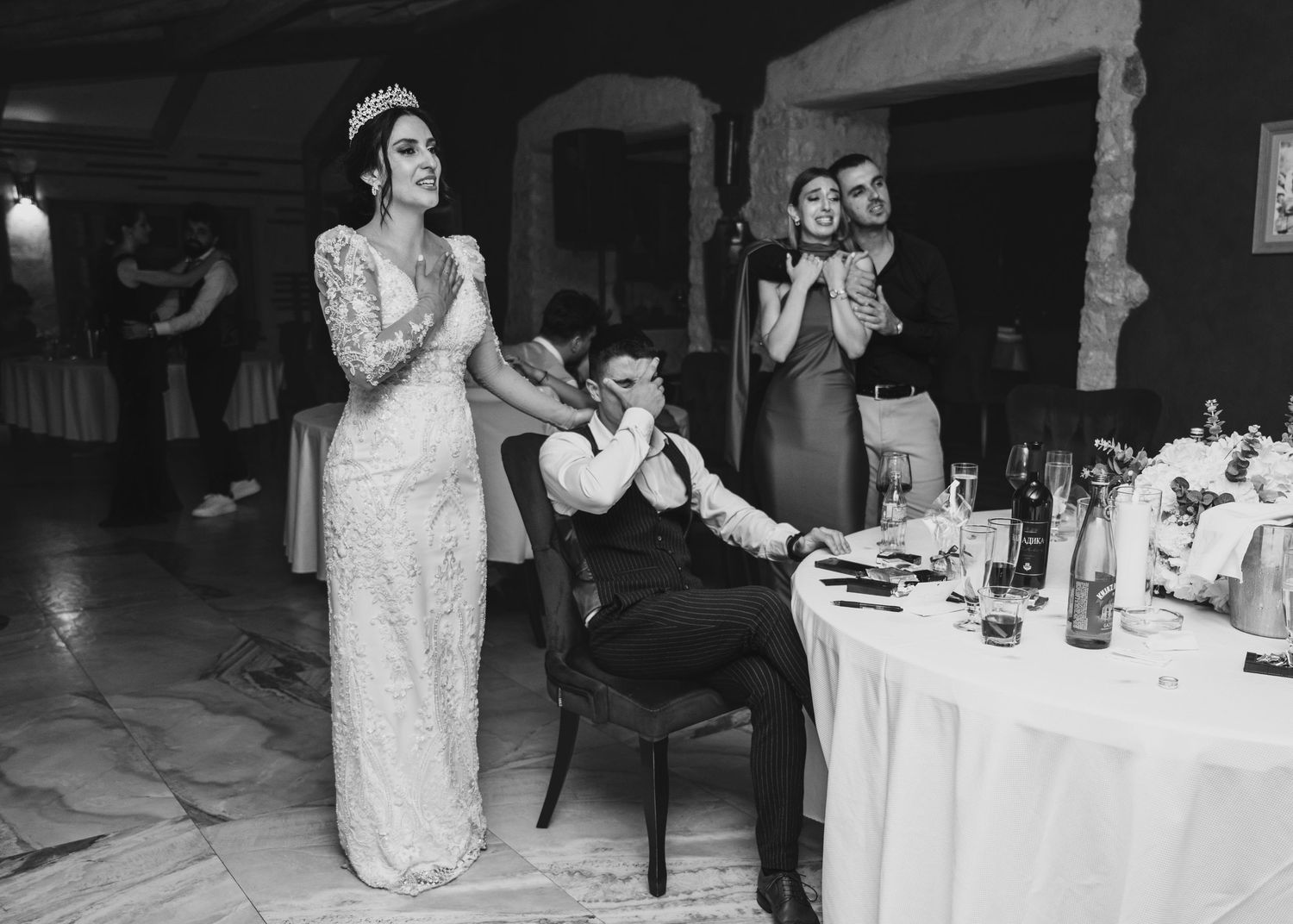 Bride in tiara comforting emotional groom at wedding reception table.