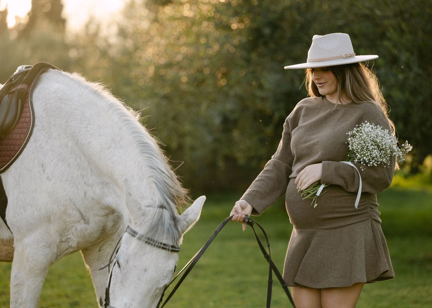 A pregnant woman in a brown dress holds flowers while standing beside a white horse at golden hour.