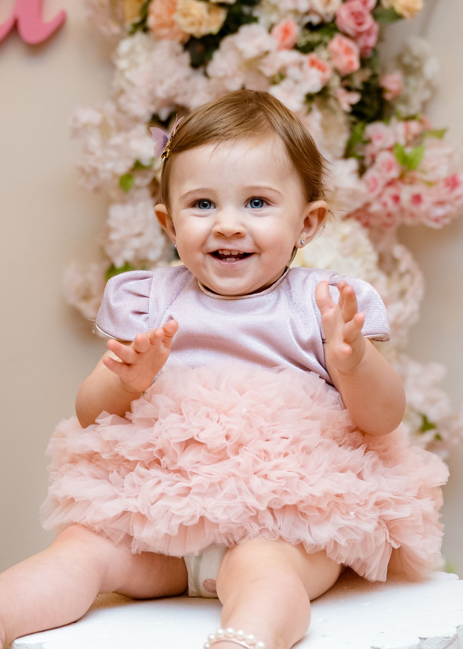 Adorable baby girl in pink ruffled dress smiling and clapping at floral backdrop.