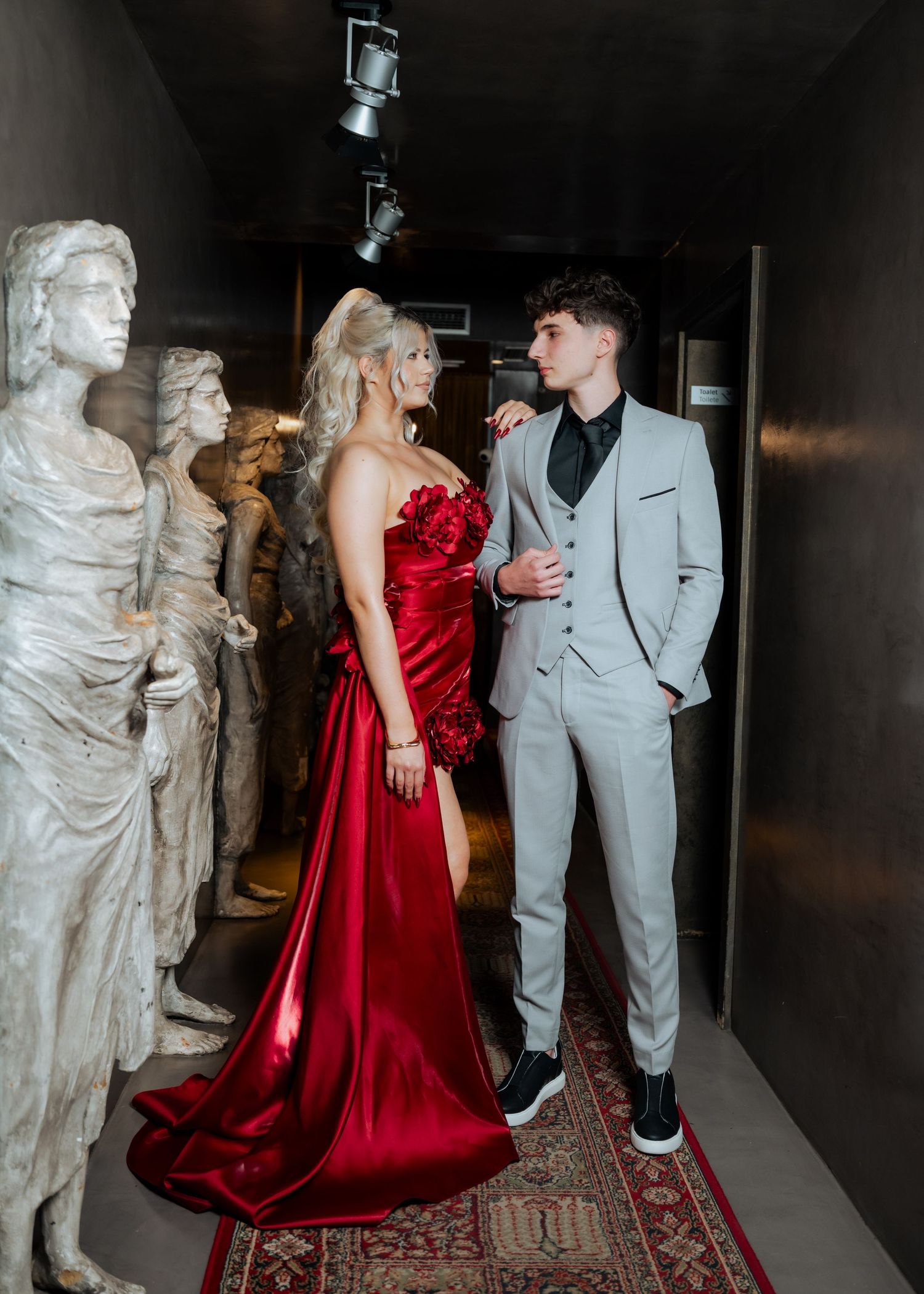 Blonde woman in red satin gown and man in gray suit posing among classical statues.