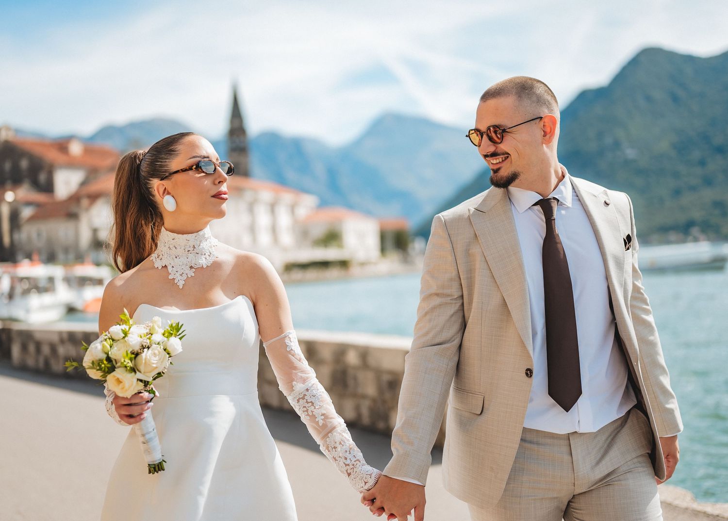 Smiling bride and groom in sunglasses walking hand in hand along Montenegro bay waterfront.