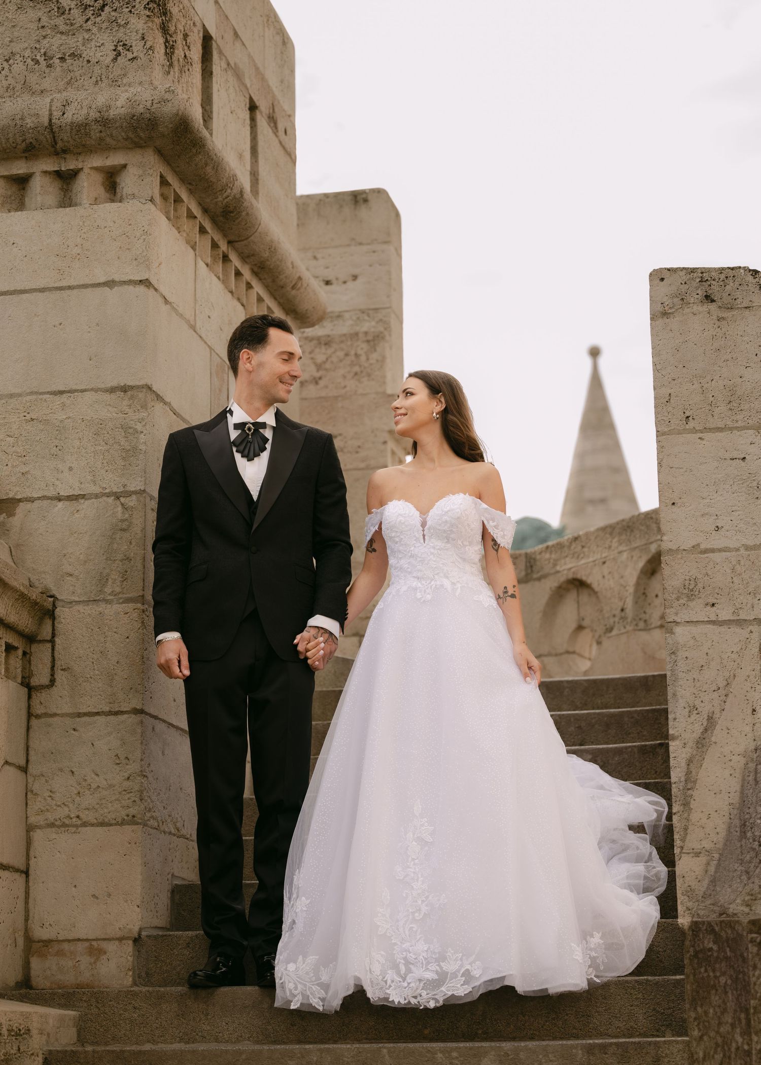 Bride in lace off-shoulder ball gown and groom in black tuxedo holding hands on castle stone steps.