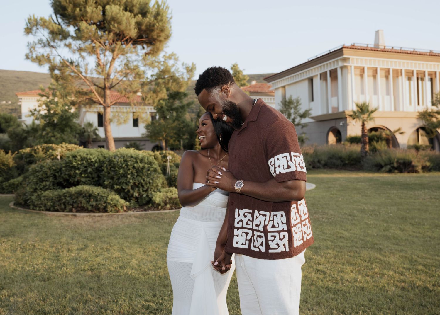 A happy engaged couple embracing and laughing together on a lush green lawn at sunset.