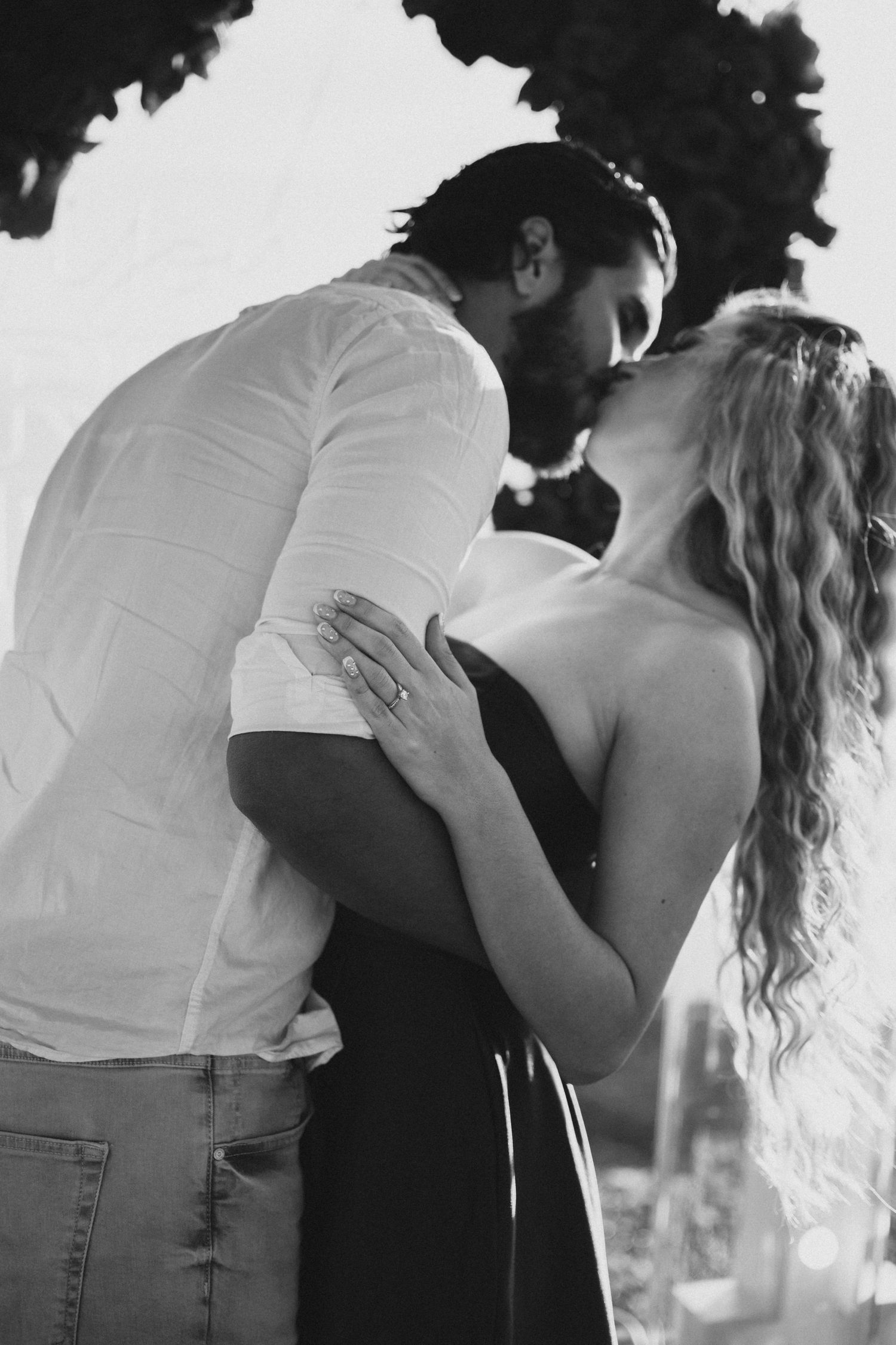 Black and white romantic photo of couple kissing outdoors after a marriage proposal with ring on her finger.