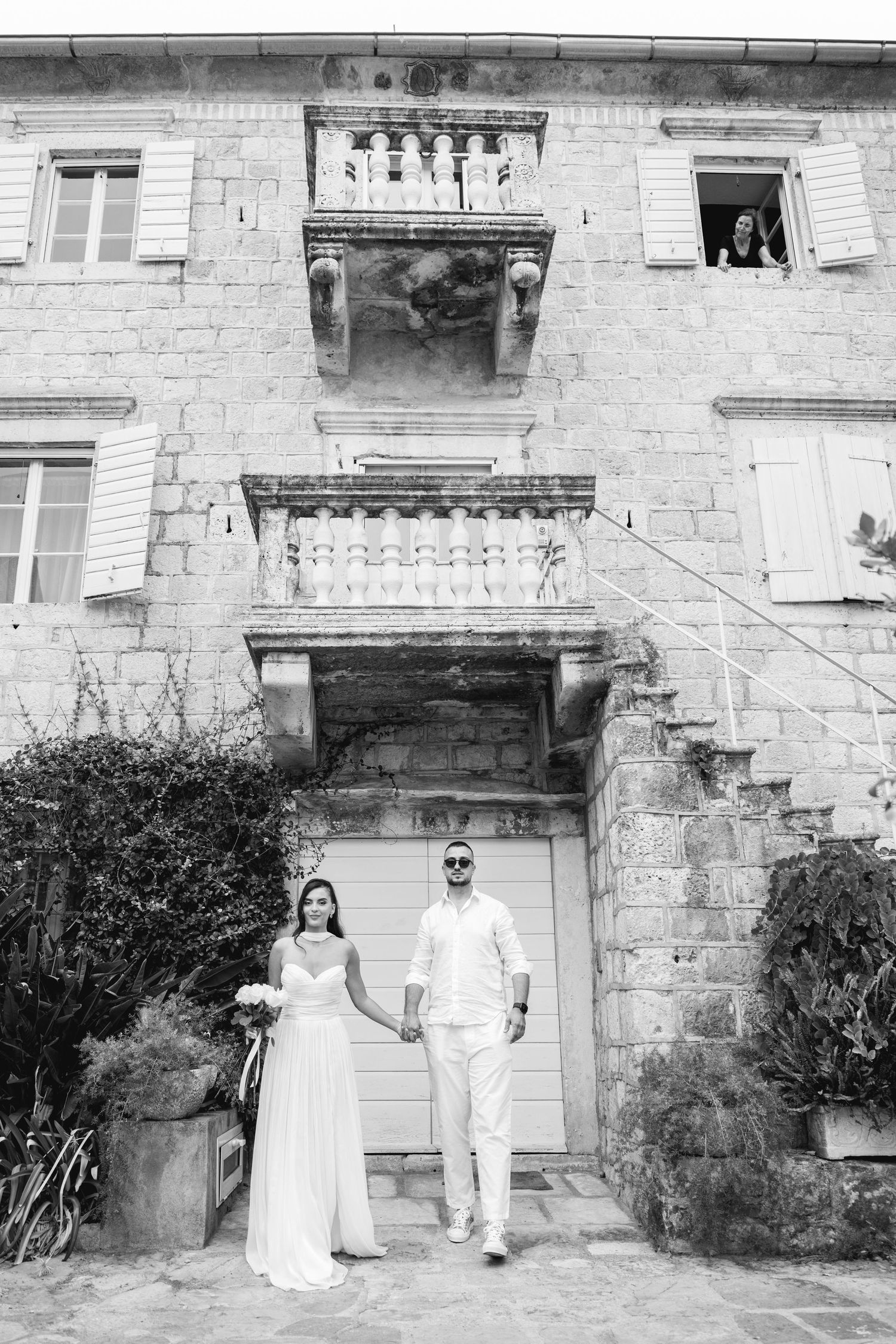 Black and white photo of bride and groom holding hands before a historic stone building in Montenegro.