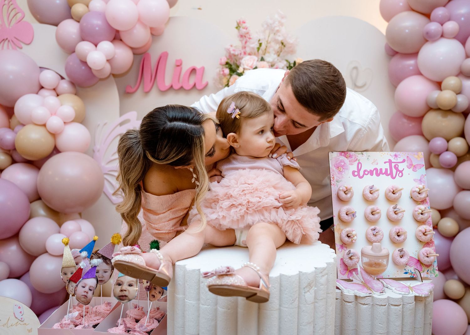Parents lovingly kissing their baby girl at her pink balloon first birthday party.