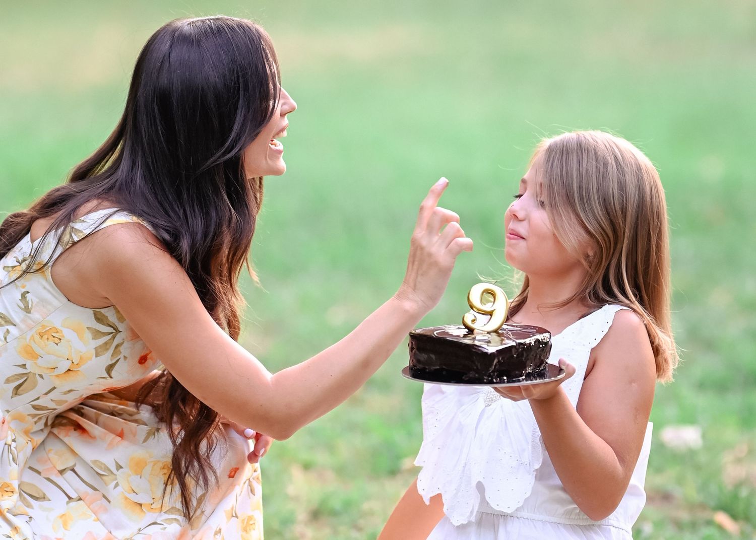 Mother and daughter sharing a birthday cake with number nine candle outdoors on green lawn.