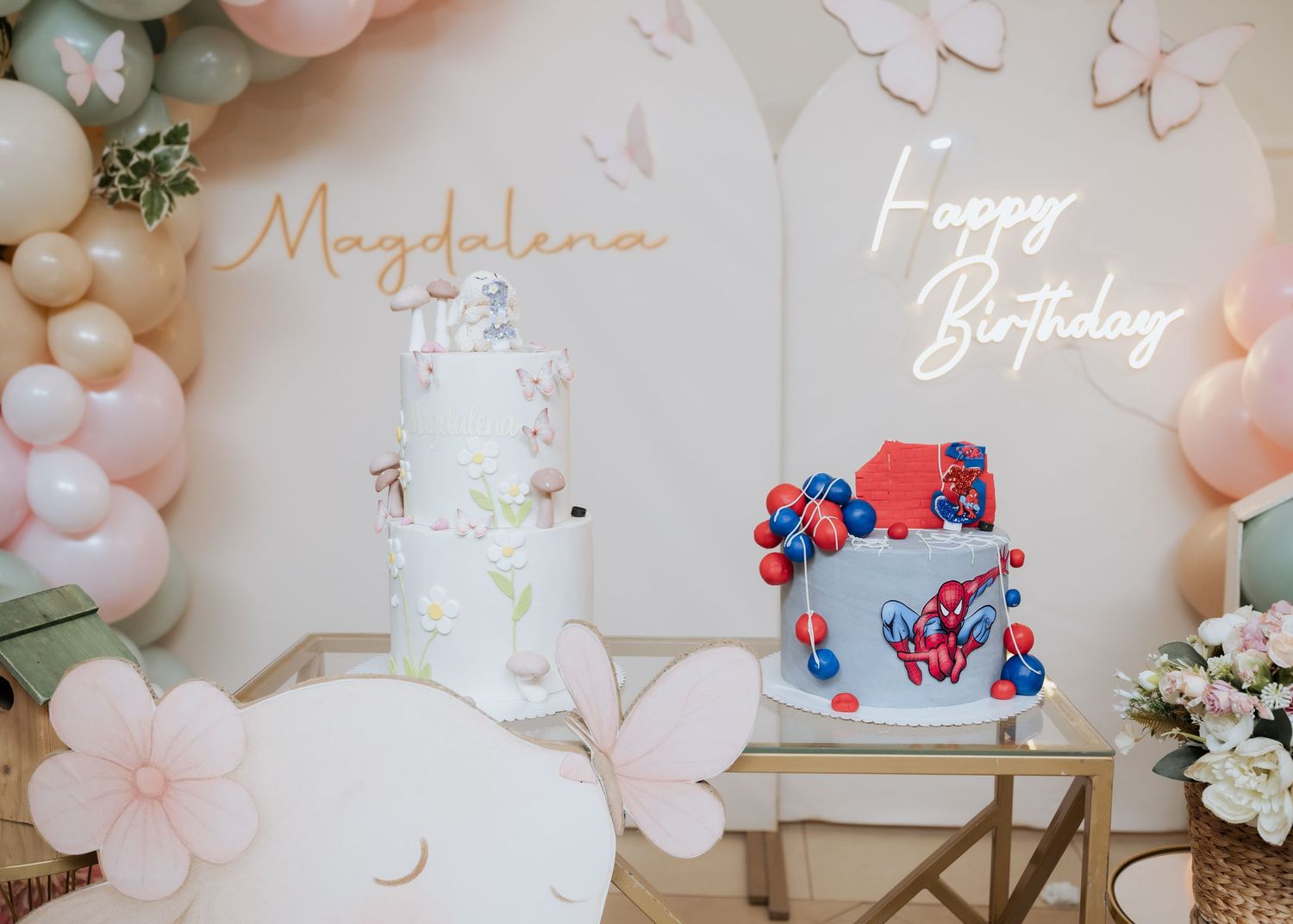Two decorated birthday cakes displayed at a party with balloon arch, one white floral and one blue with red butterflies.