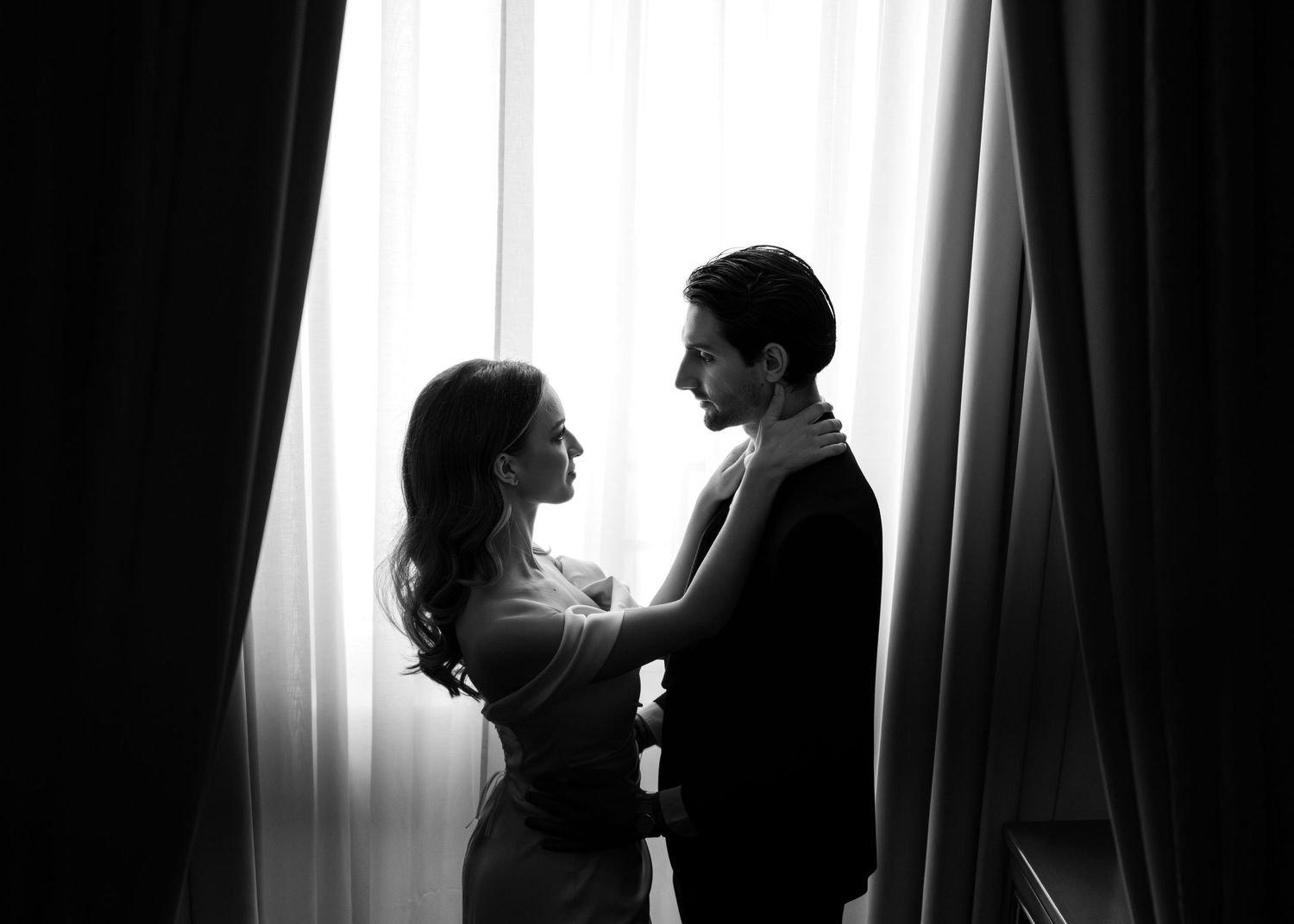 Black and white romantic silhouette of bride and groom embracing by sheer curtained window.