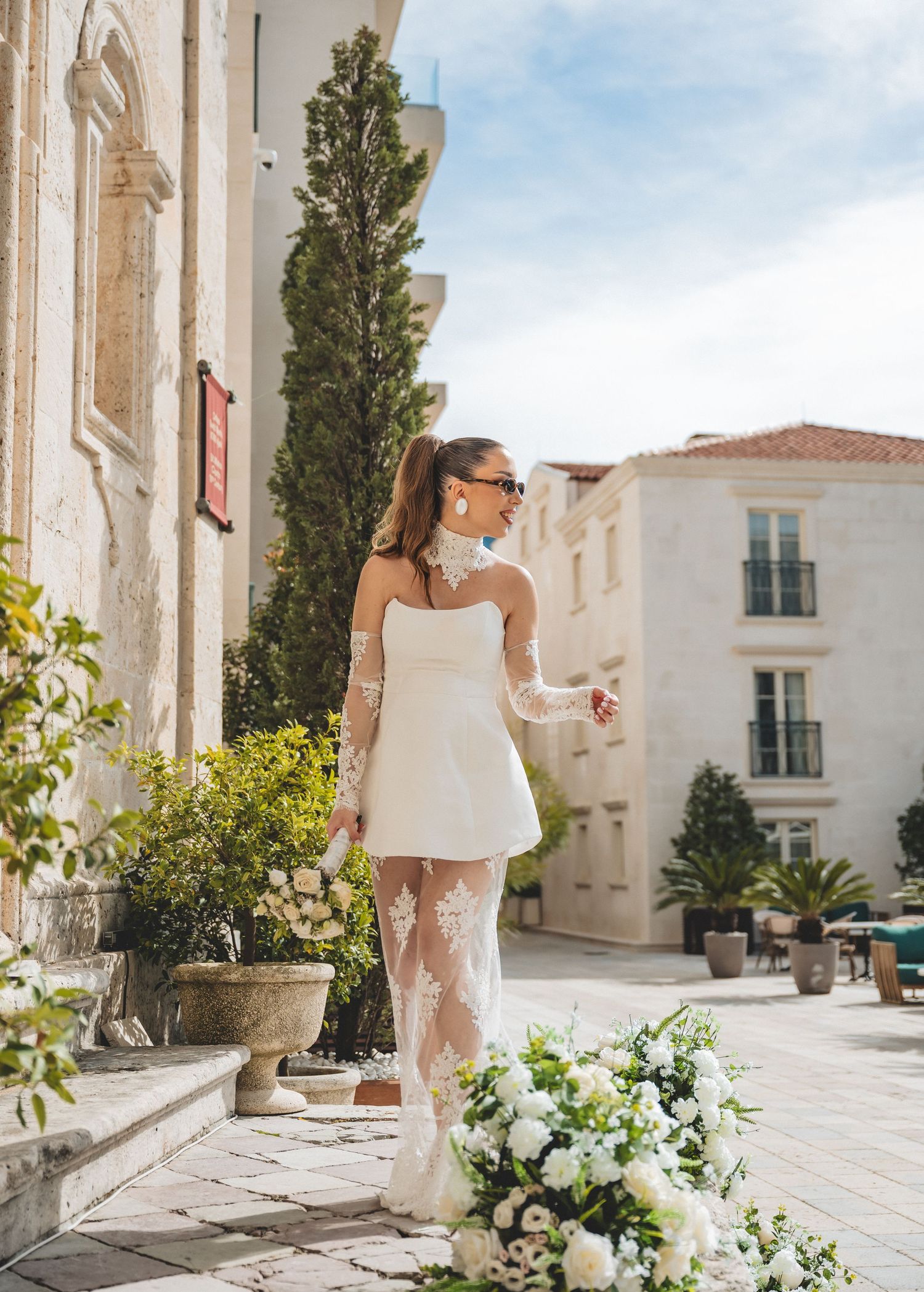 Elegant bride in short white dress with lace overlay posing in sunny Mediterranean courtyard.