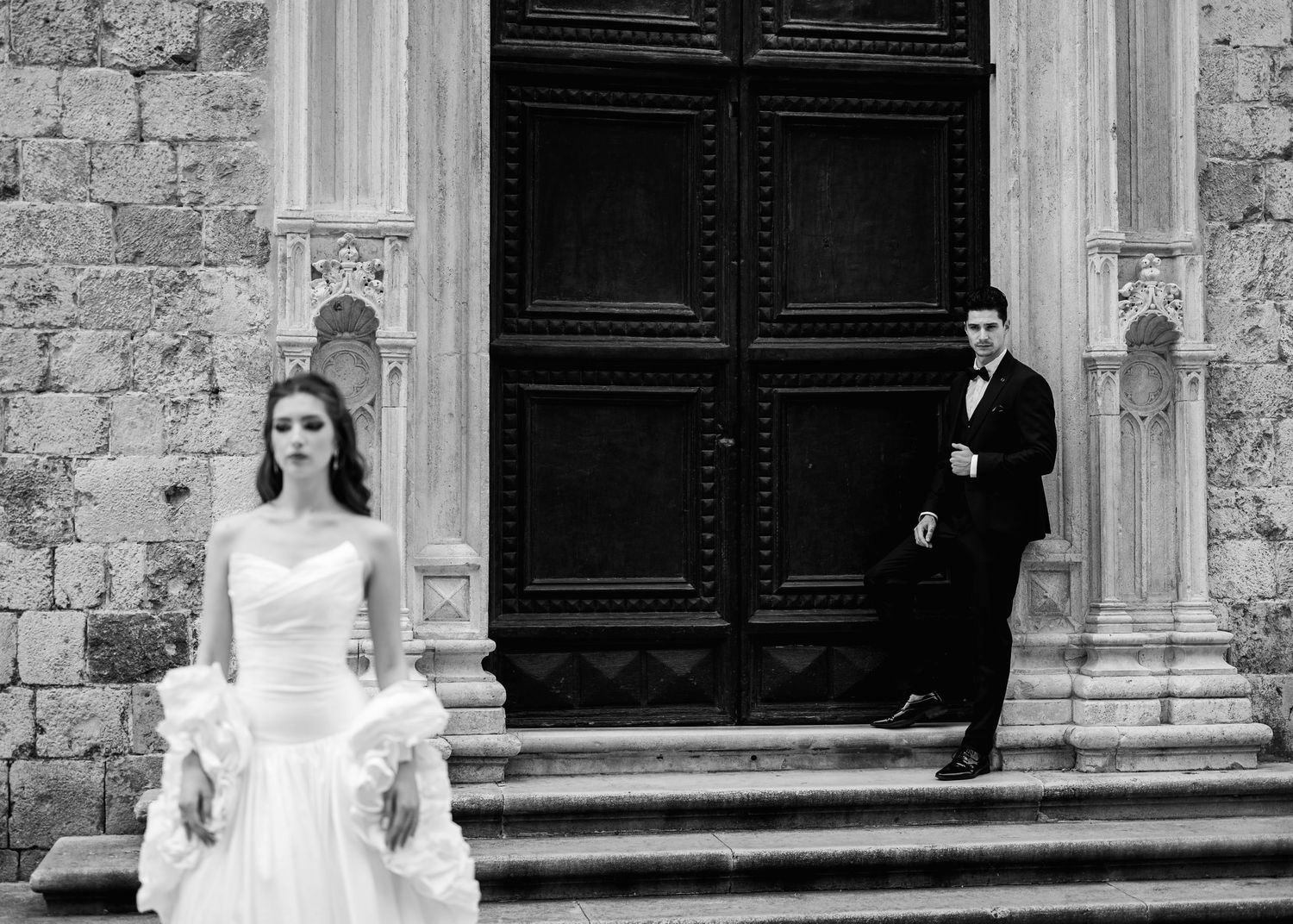 Black and white photo of bride and groom posing separately on steps before massive ornate wooden doors.