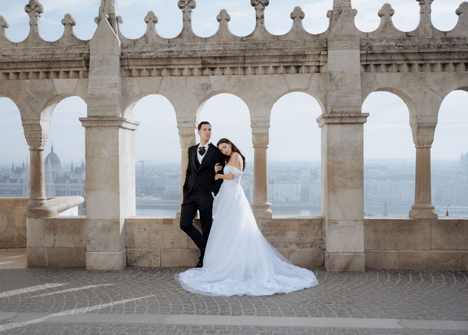 Wedding couple standing beneath ornate stone arches with panoramic Budapest city views behind them.
