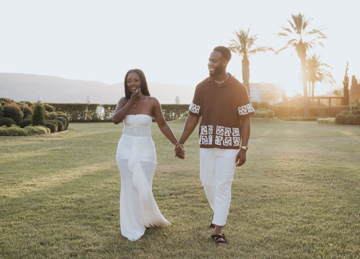 A smiling engaged couple holding hands and walking together in a sunlit outdoor garden setting.