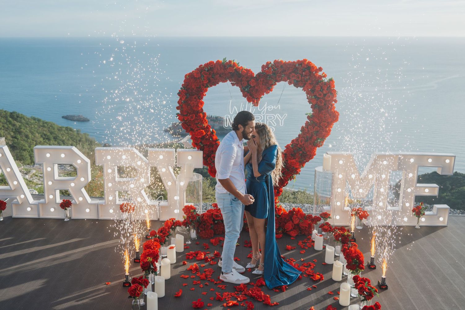 Couple kissing under red rose heart arch with 'Marry Me' marquee letters and sparklers by the ocean.