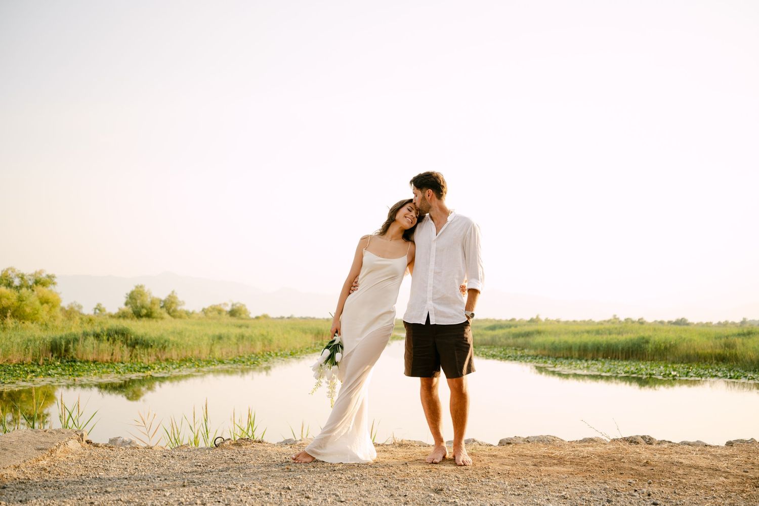 Couple in white wedding attire standing by a serene lake with lush green fields at golden hour.