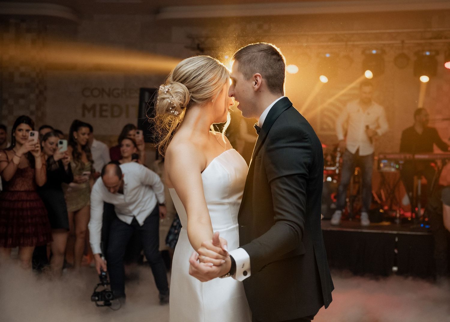 Bride and groom share romantic first dance with fog effects and guests photographing at wedding reception.