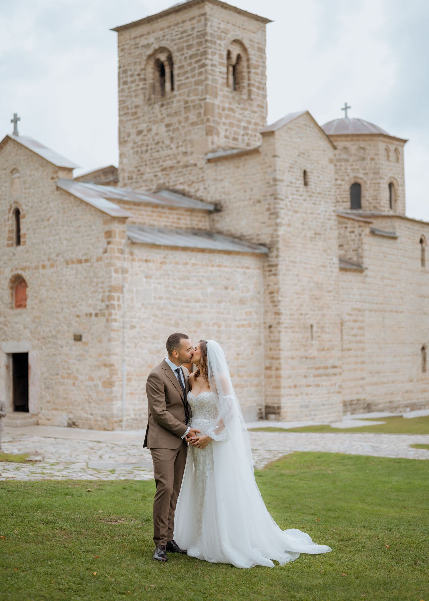 Newlyweds kissing on green lawn in front of historic stone medieval church on wedding day.