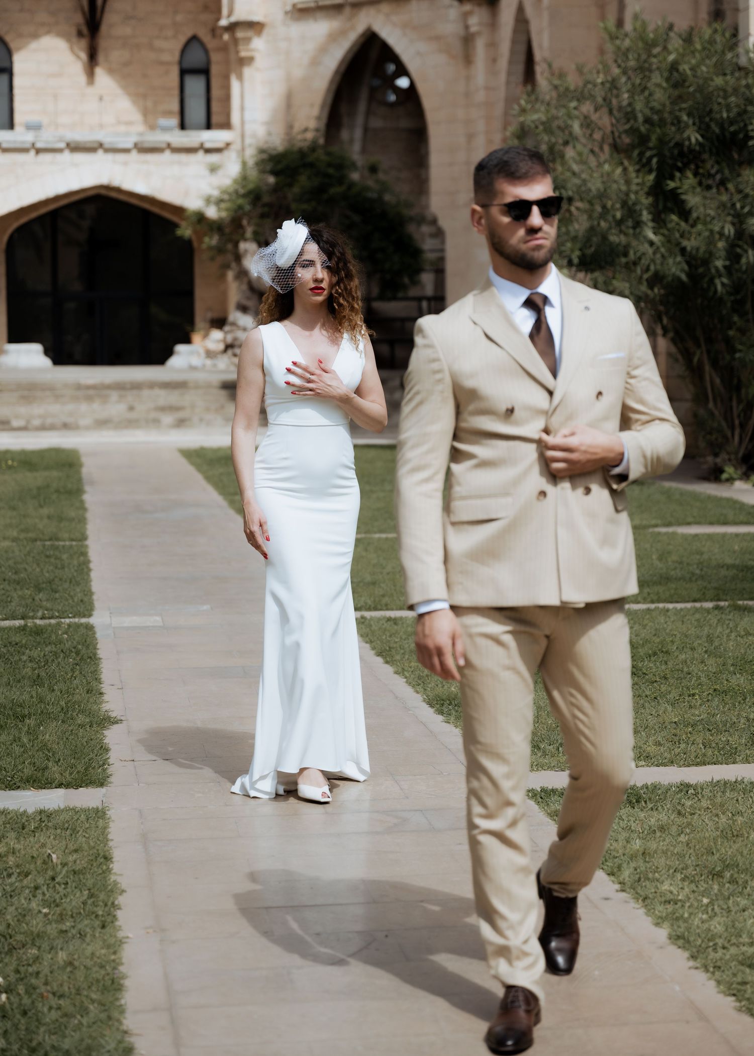 Stylish bride and groom pose separately in beige suit and white gown at Gothic ruins.