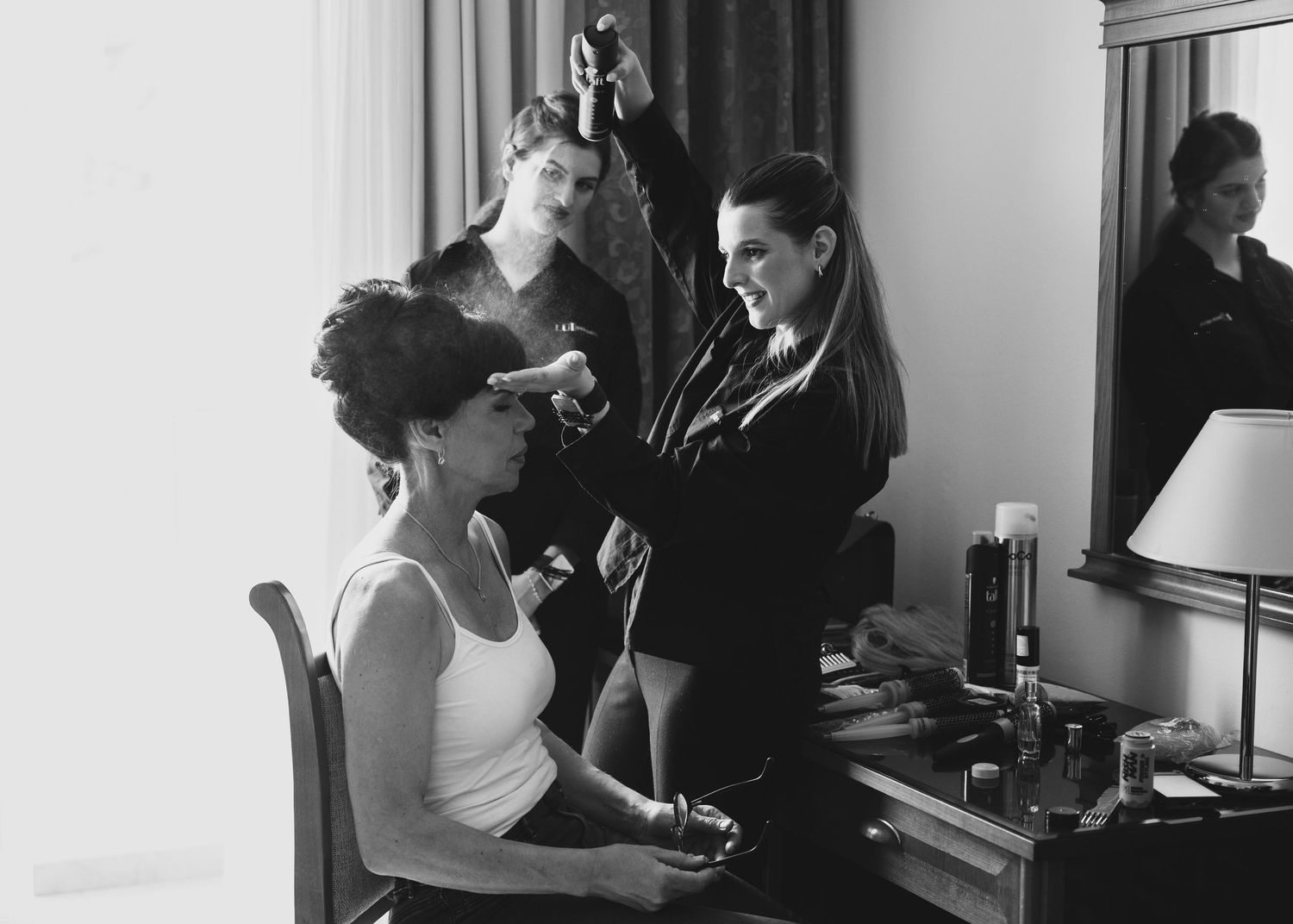 Black and white photo of a bride getting her hair styled by a professional stylist in a hotel room.