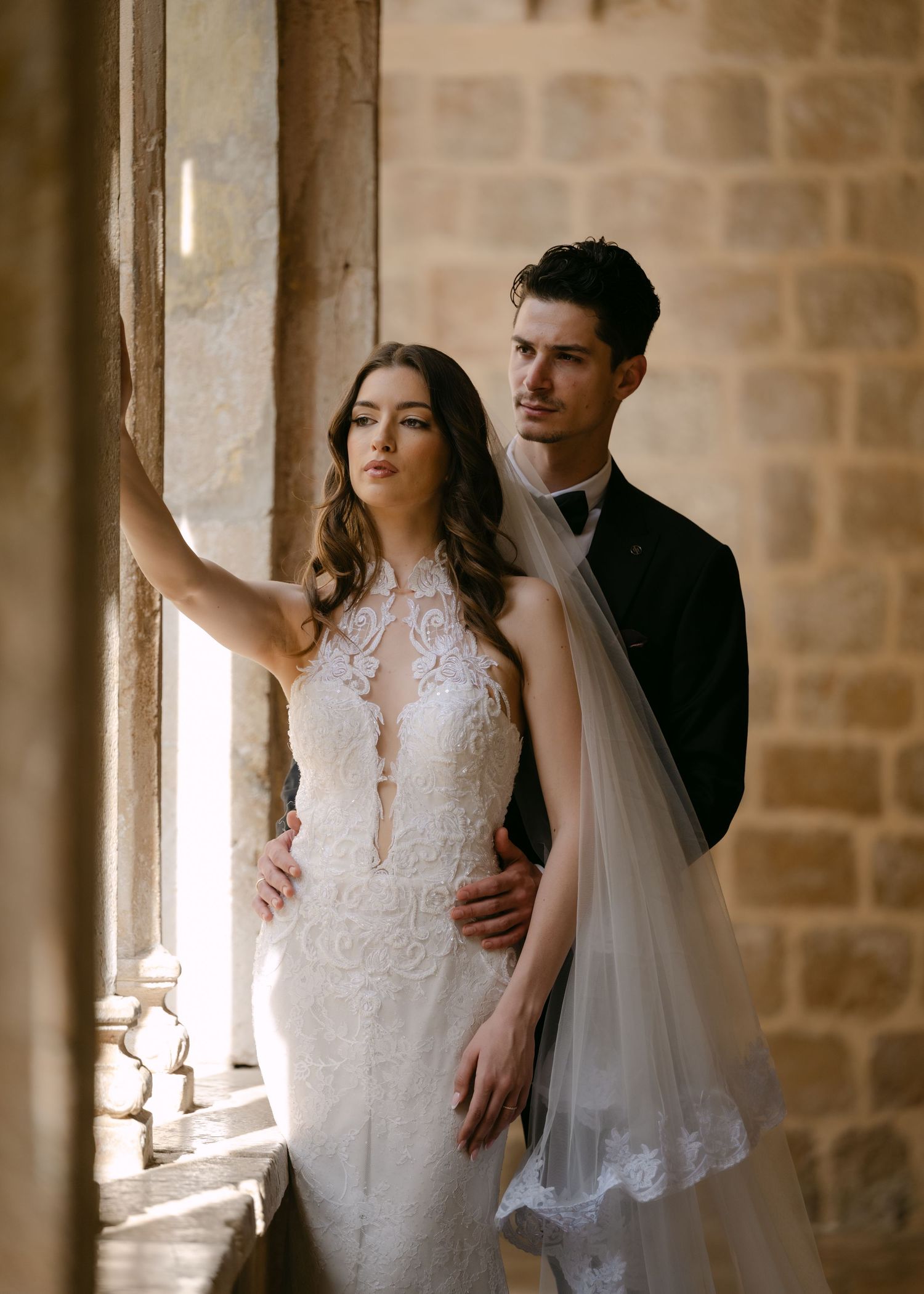 Bride in lace halter wedding gown and groom in black tuxedo posing by stone archway window.