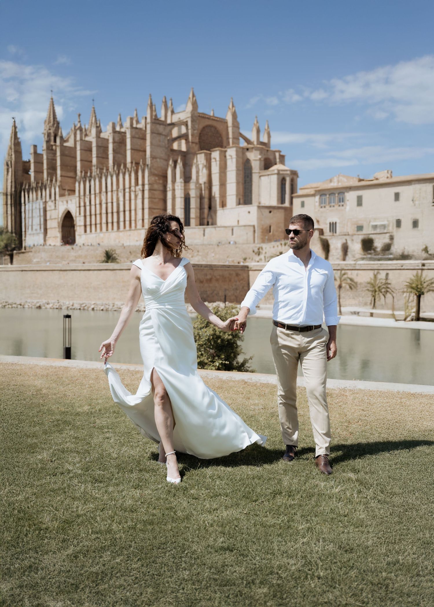 Bride and groom walk hand-in-hand on green lawn with stunning Gothic cathedral backdrop.