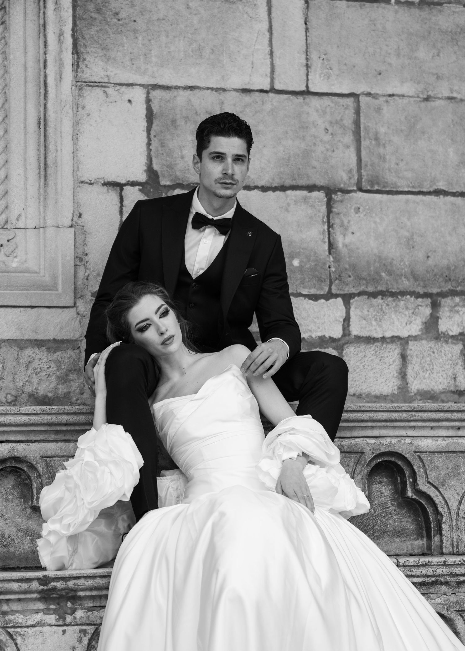 Classic black and white wedding portrait of bride in ruffled ball gown and groom in tuxedo against a stone wall.