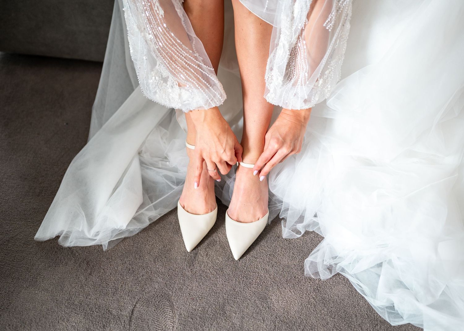 Bride in lace wedding dress fastening ankle strap on pointed white bridal heels.