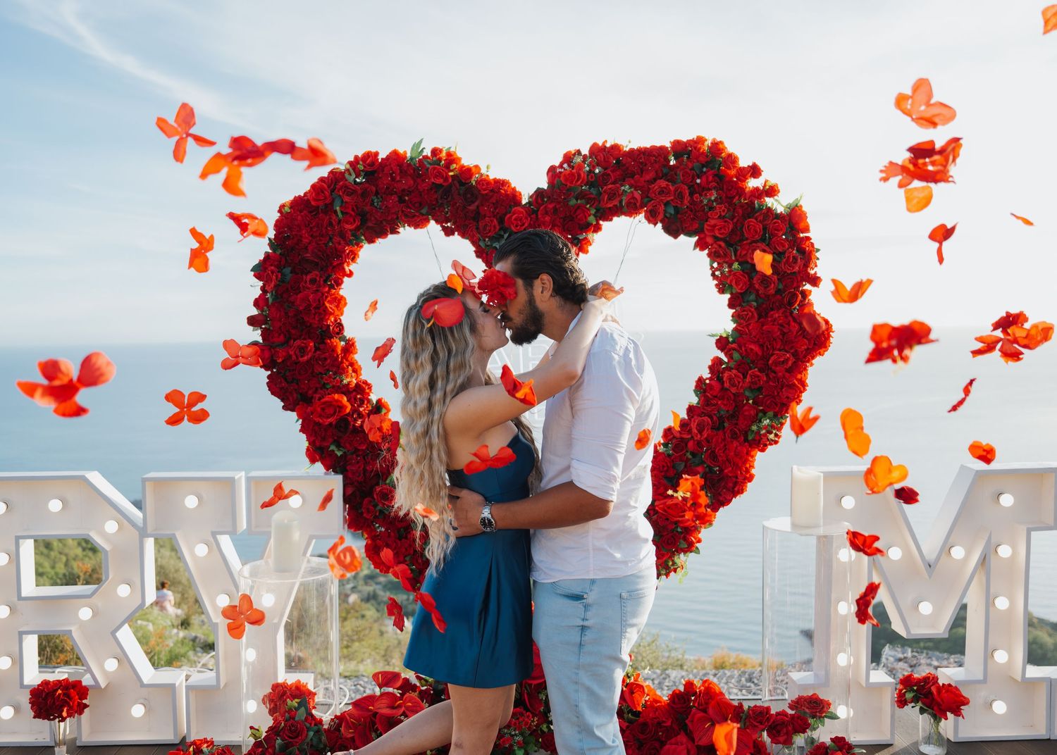Couple kissing in front of a large red rose heart arch with 'Marry Me' letters and rose petals falling outdoors.