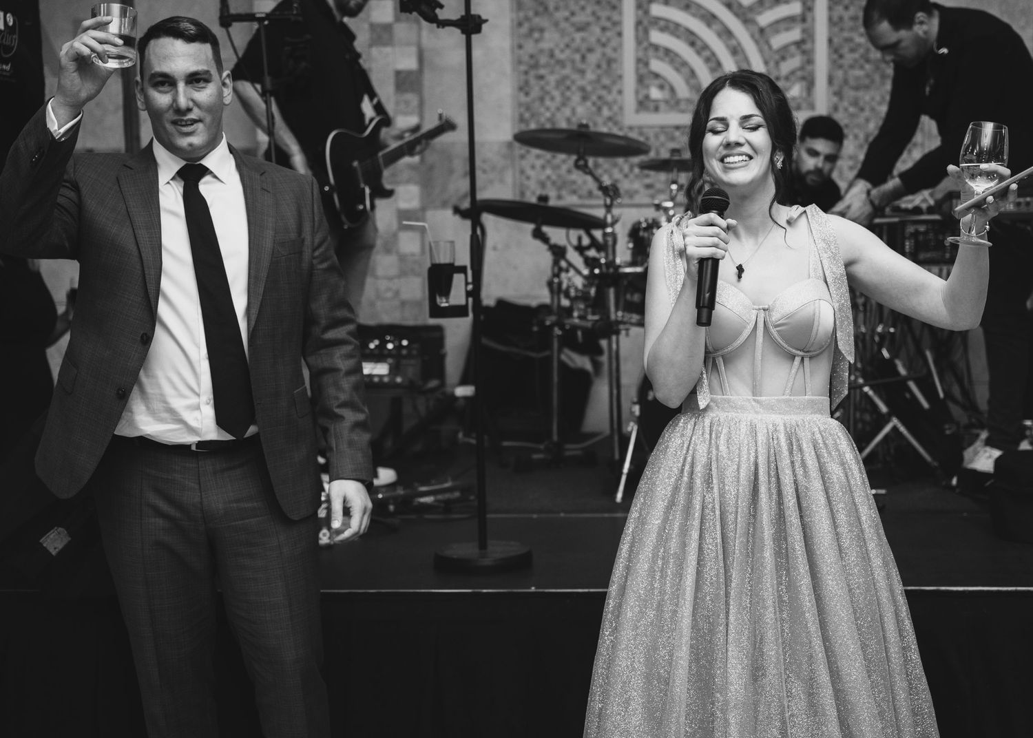 Wedding hosts raising glasses on stage with live band performing at elegant wedding reception.