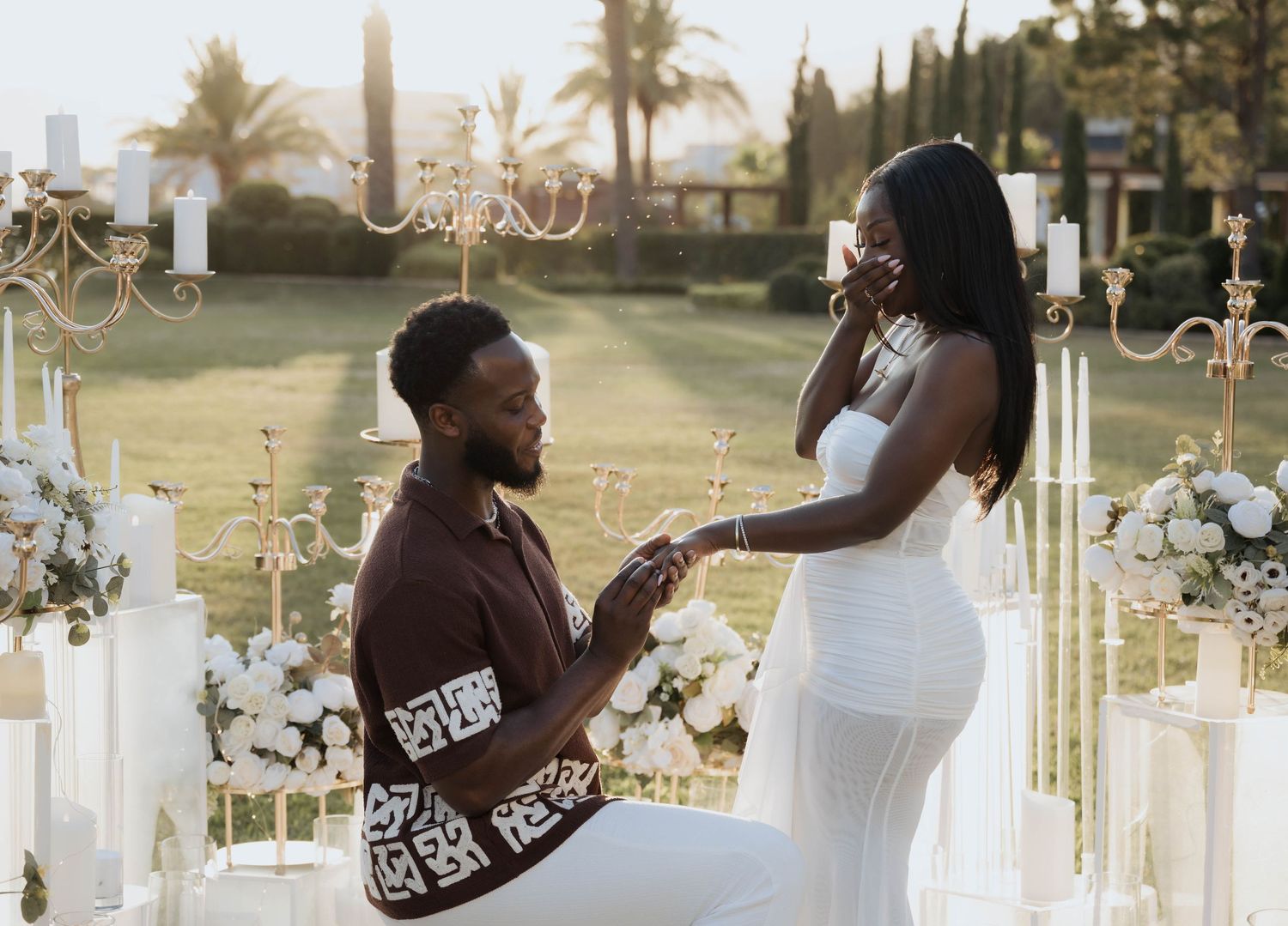 A man kneeling to propose to a woman in a white dress at a candlelit outdoor ceremony.