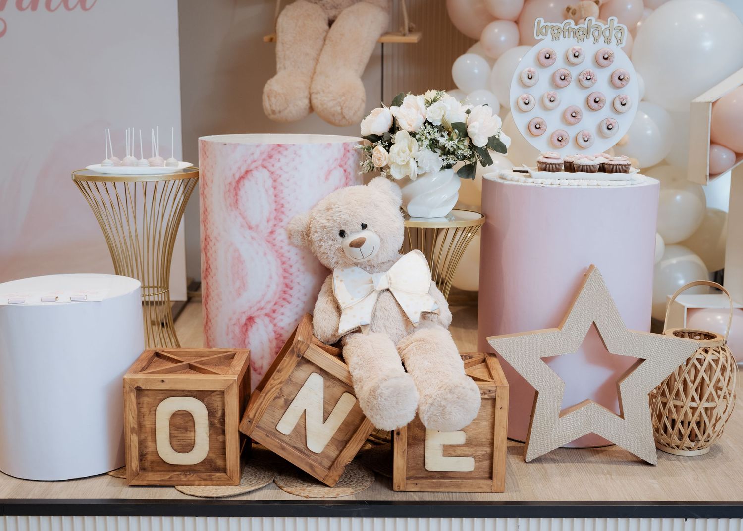 Teddy bear sitting on wooden ONE blocks surrounded by pink party decorations and flowers.