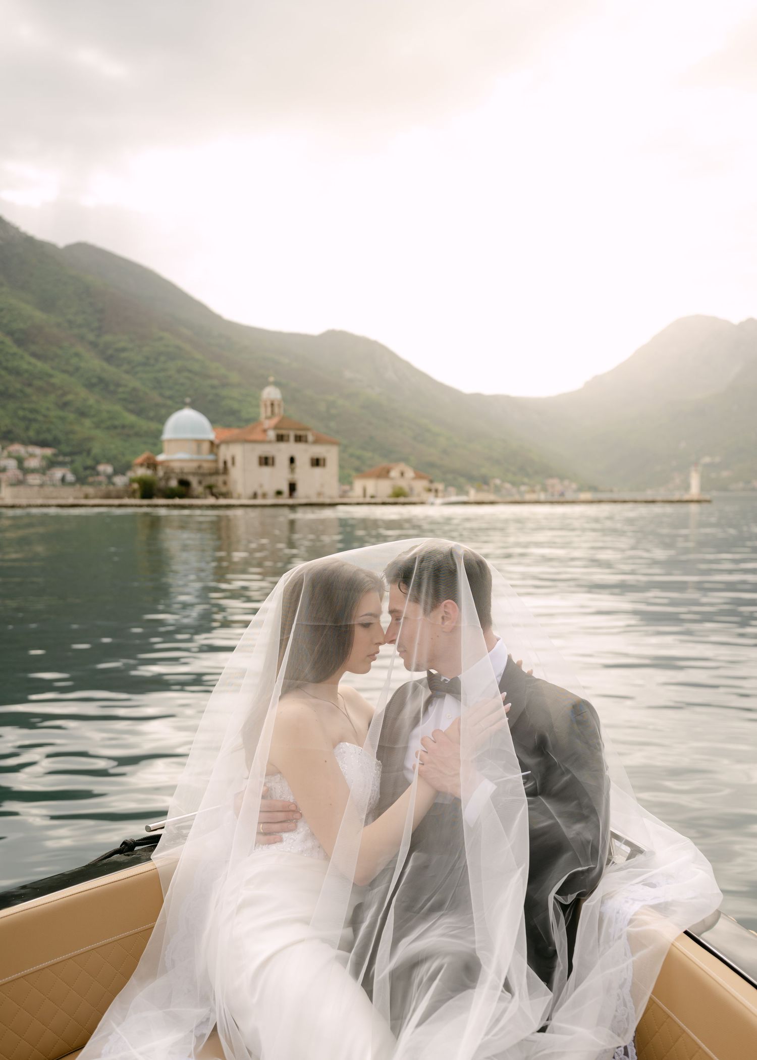 Bride and groom share intimate moment under veil on boat with scenic bay backdrop.