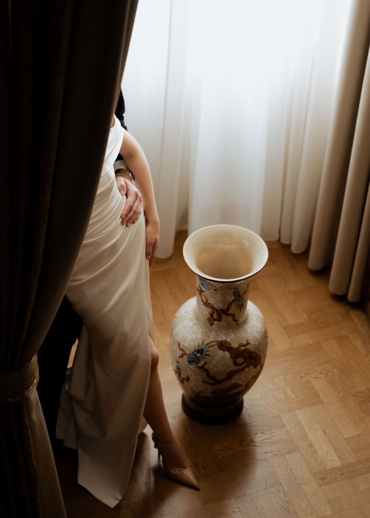 Elegant bride in a white gown stands by sheer curtains next to a large ornate ceramic vase on a parquet floor.