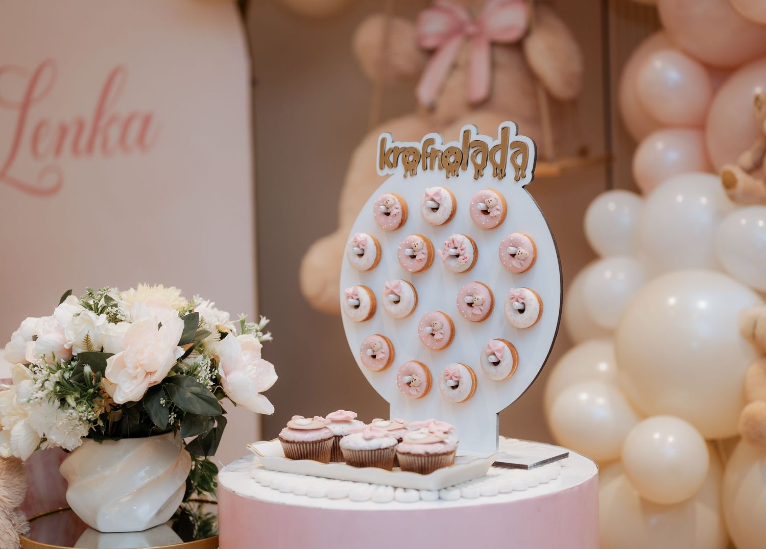 Donut wall display on a white cake with pink frosting donuts and Lenka sign at a birthday party.
