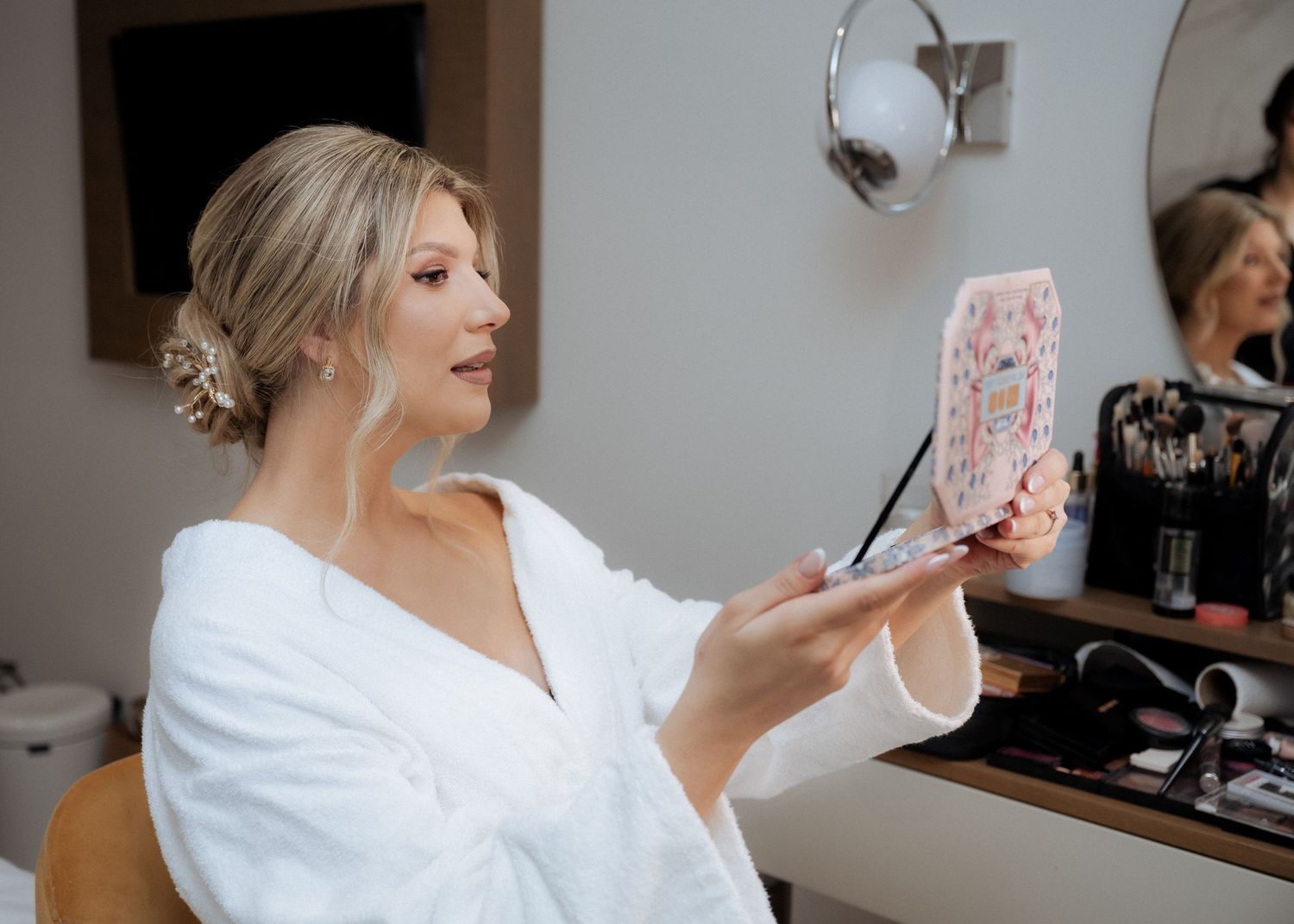 A blonde bride in a white robe checking her makeup in a handheld mirror at a vanity.